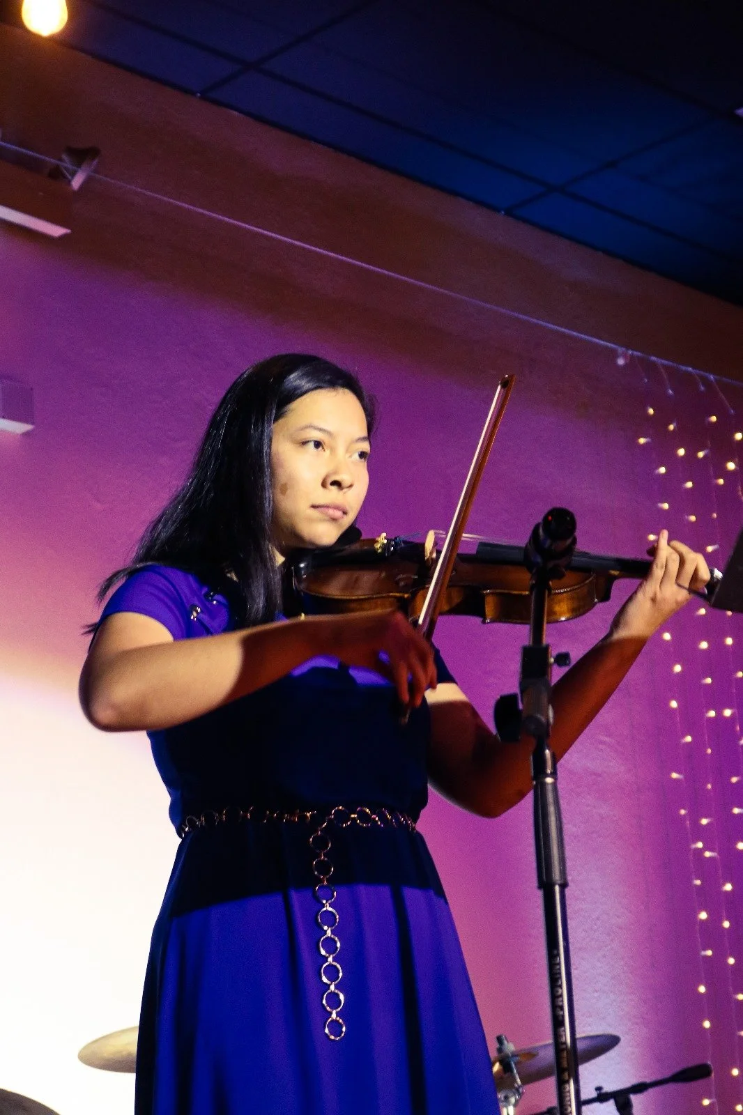 A young woman playing the violin on stage, illuminated by purple and pink lighting, with string lights in the background.