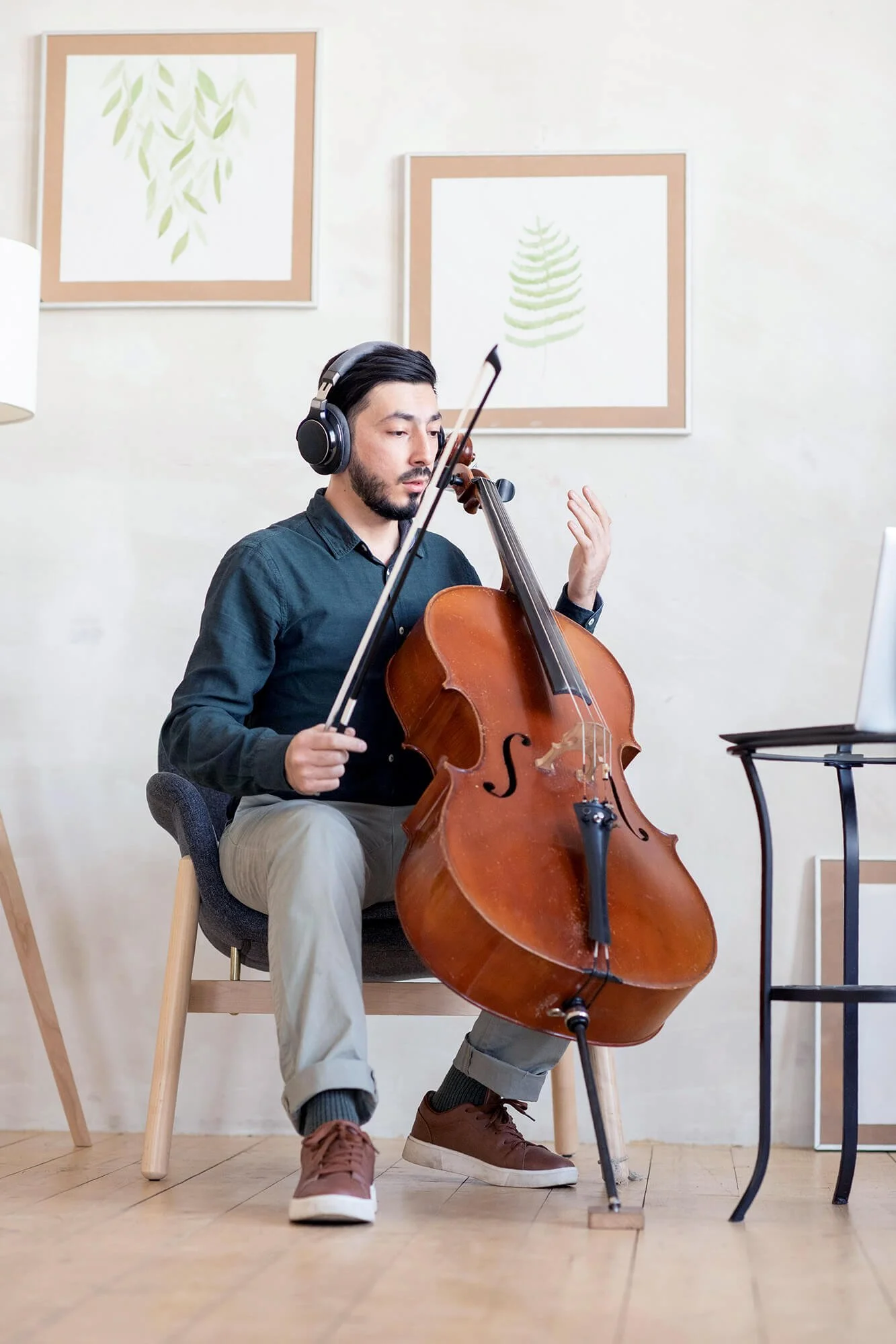 A man sitting on a chair playing a cello while wearing headphones, in a room decorated with framed botanical artwork on the wall.