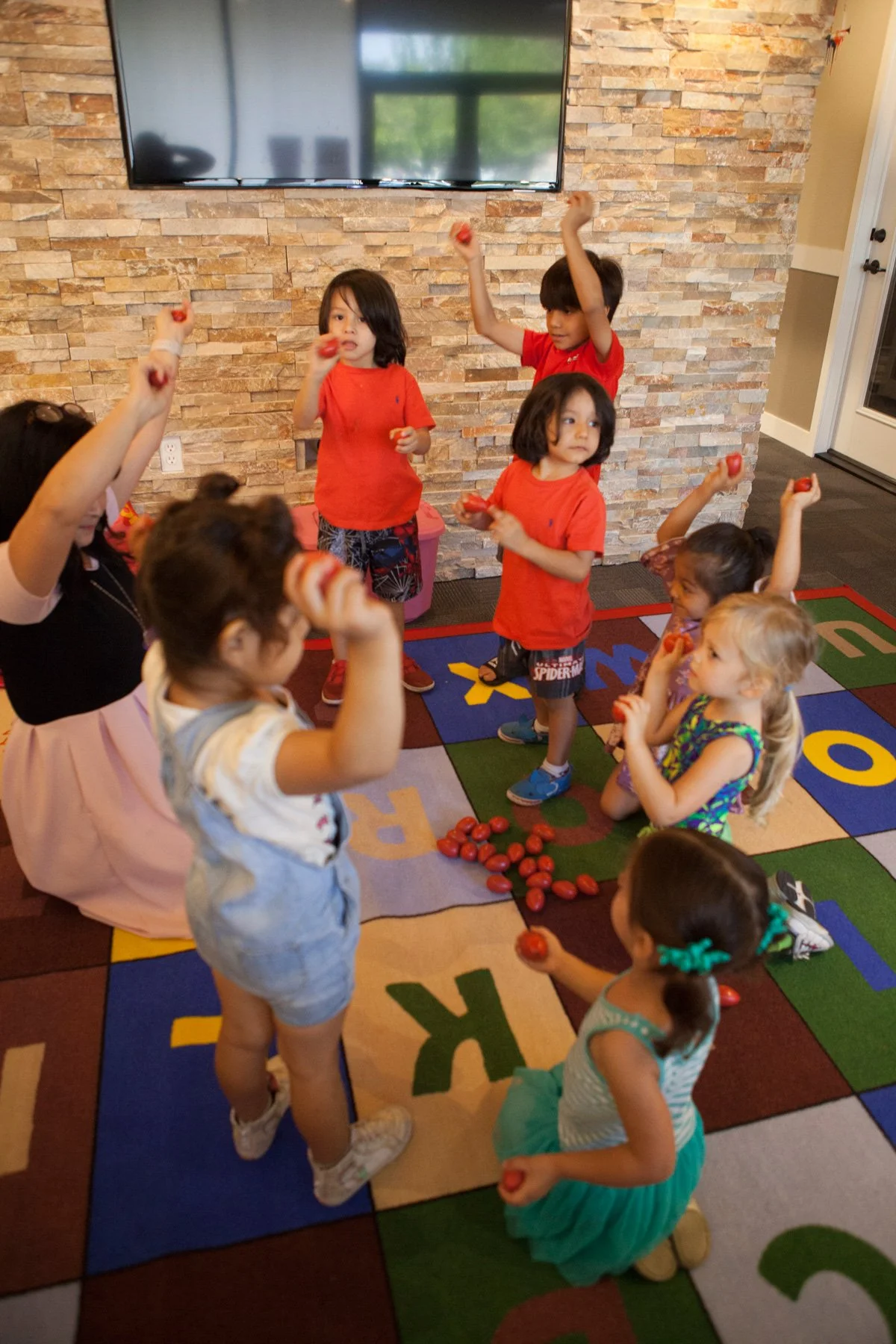 Children and a woman in a room with a stone wall, gathered on a colorful alphabet mat, holding and eating cherry tomatoes.