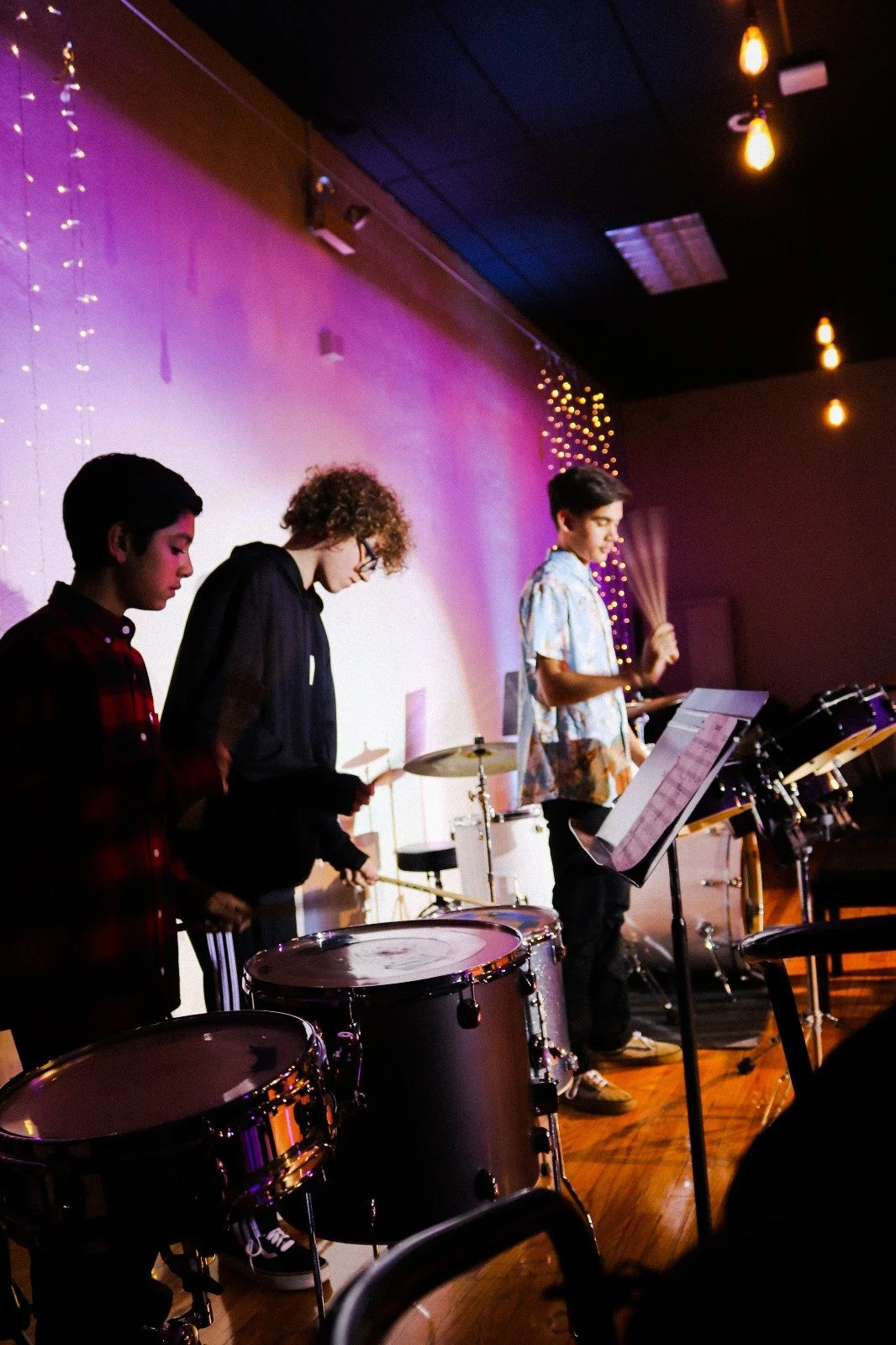 Three young musicians playing drums on stage in a dimly lit room with colorful lighting and decorative string lights.