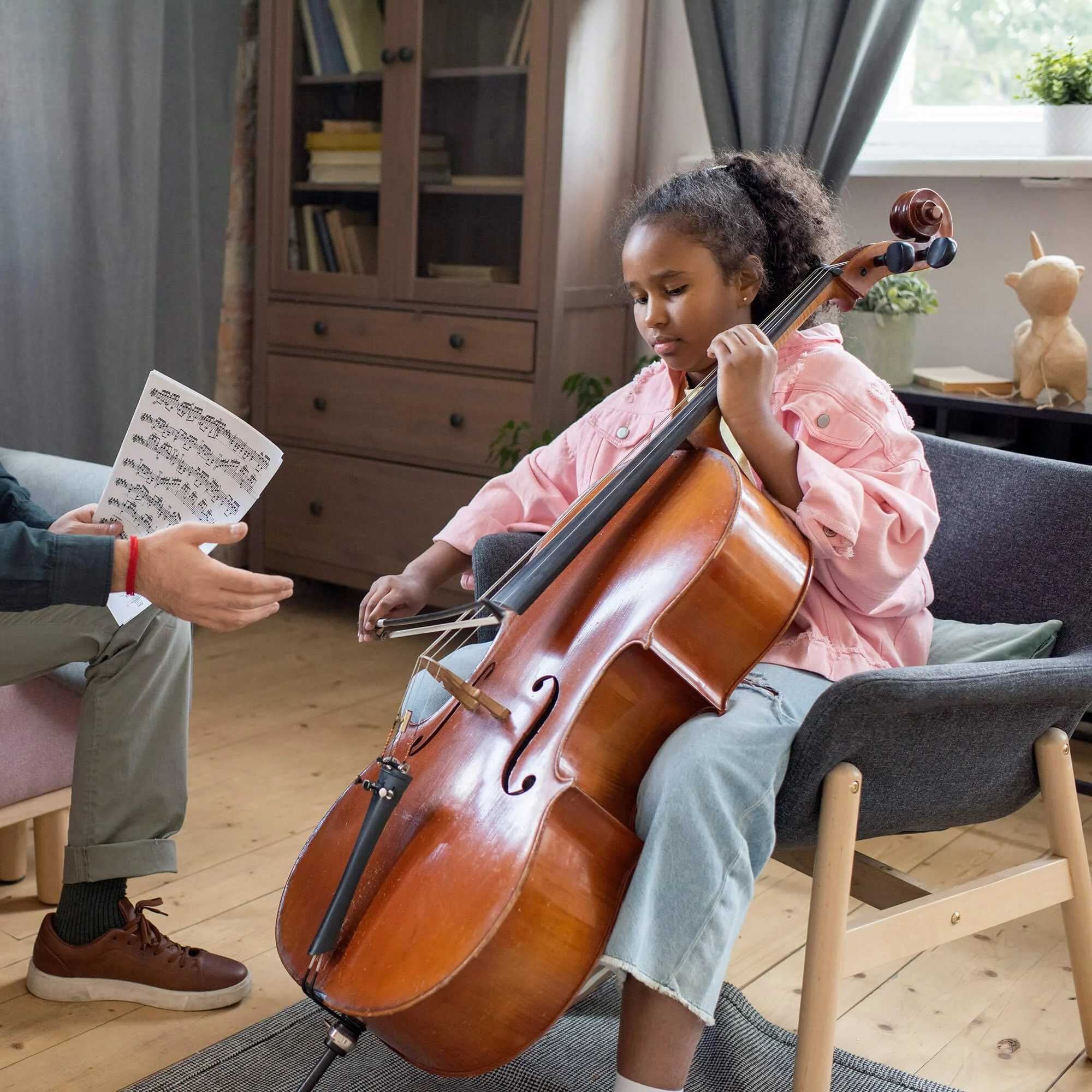A young girl playing the cello during a music lesson with an instructor holding sheet music.
