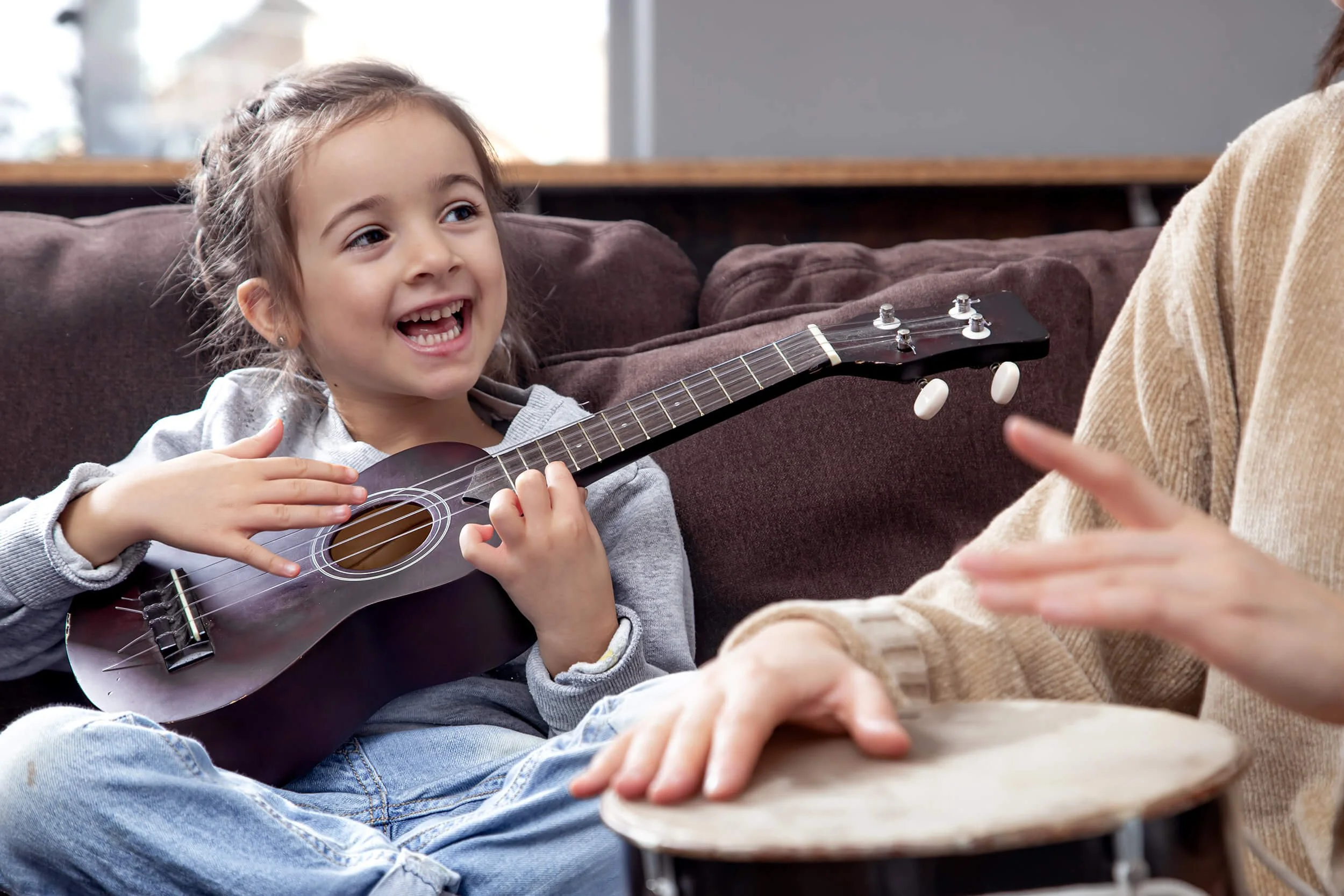 A young girl with brown hair and a gray hoodie, sitting on a dark-colored couch, smiling and playing a small black ukulele while an adult plays a drum with their hand.