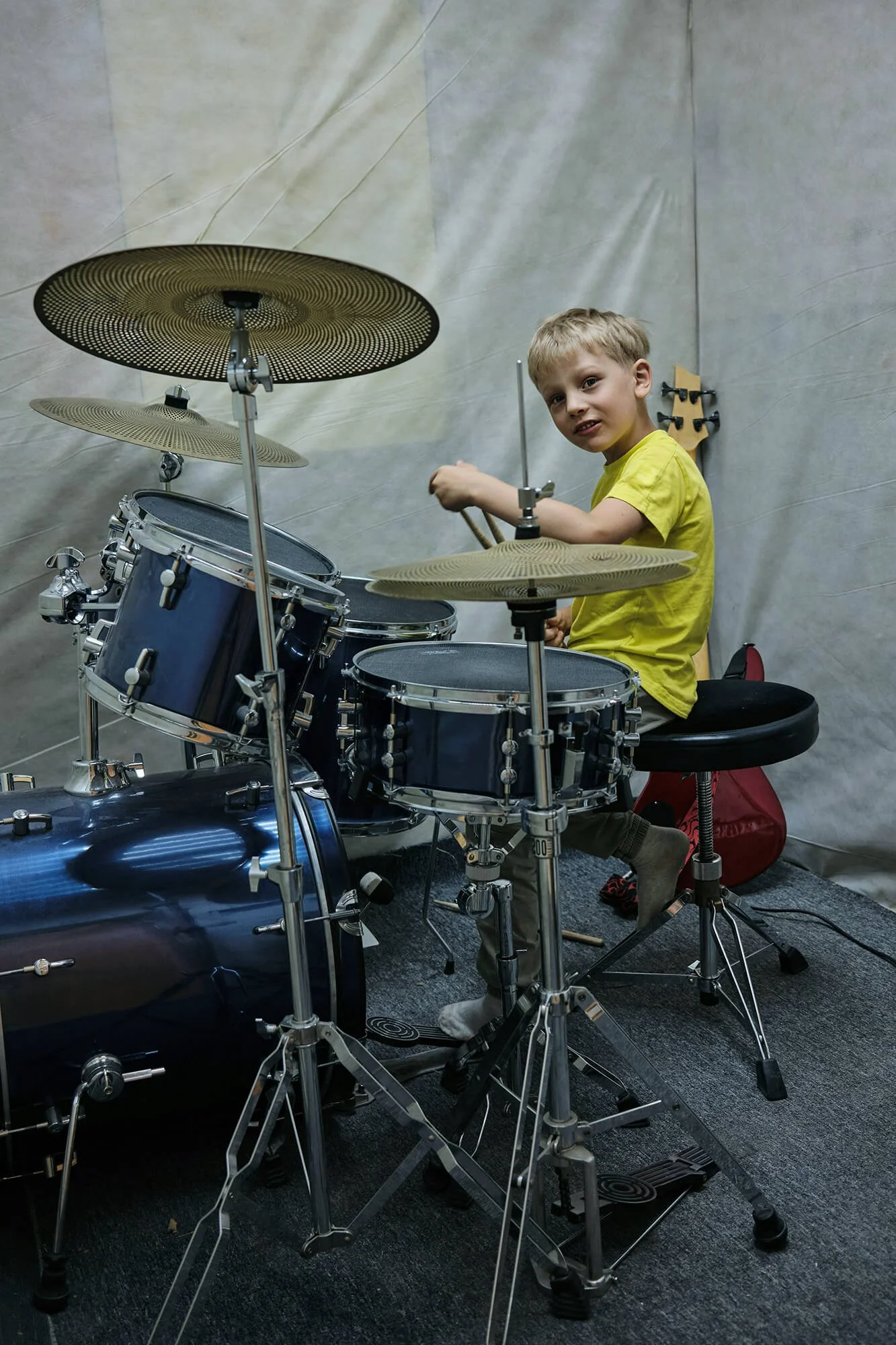 A young boy in a yellow t-shirt sitting at a drum set, playing drums in a music practice room.
