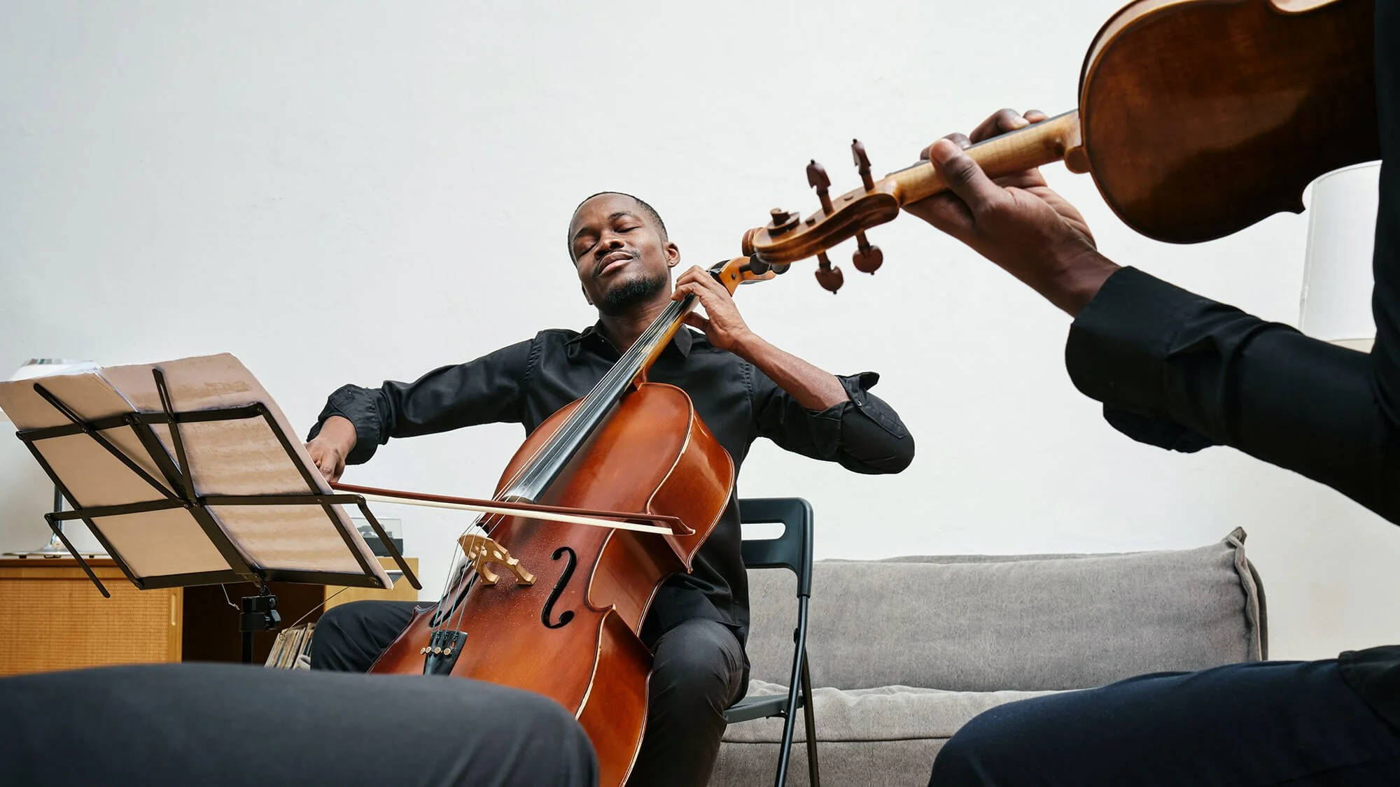 Two men playing classical guitar and cello in a room with a white wall, one sitting on a chair, the other grabbing the guitar