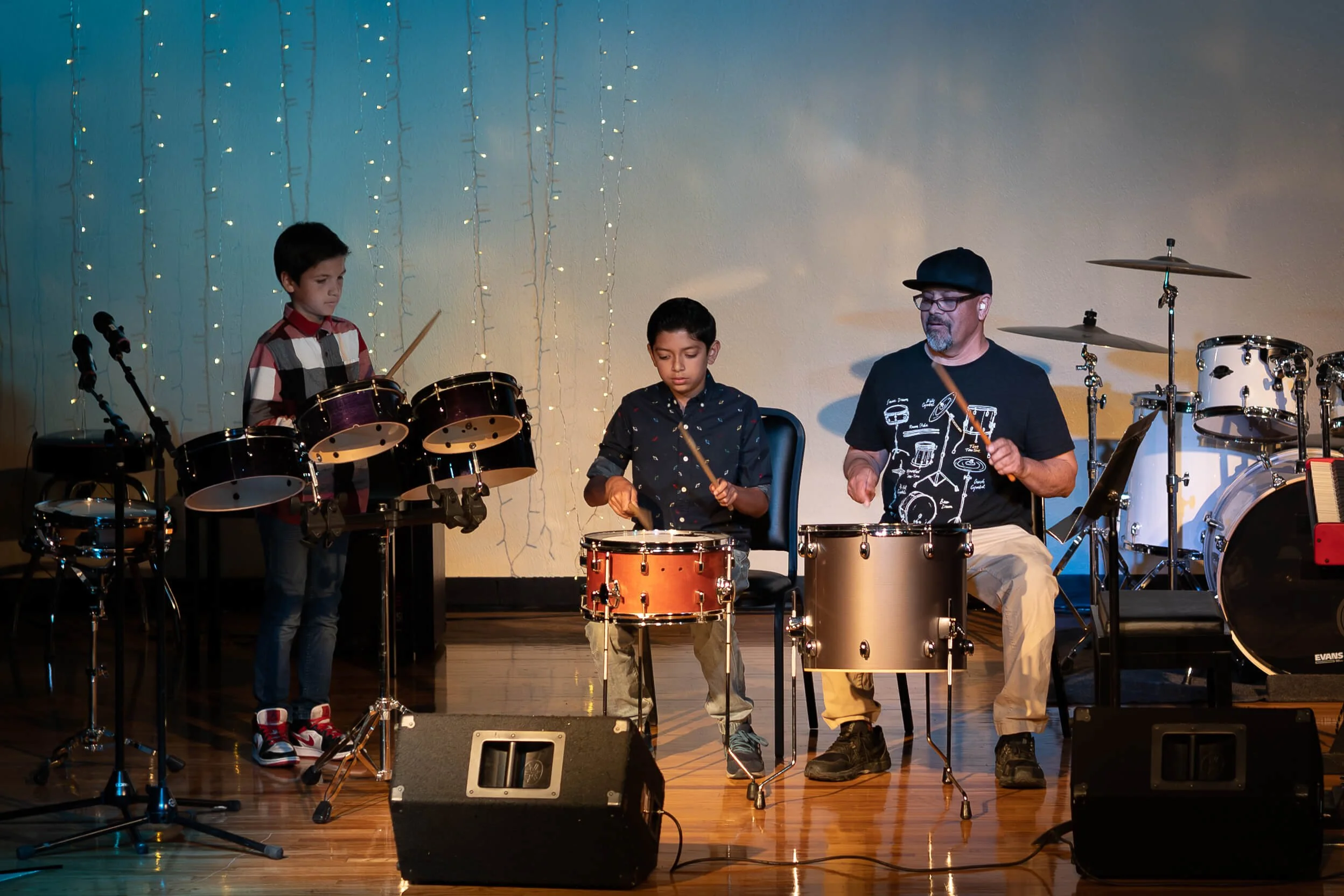 Three people playing drums on stage during a performance, with a backdrop decorated with string lights.