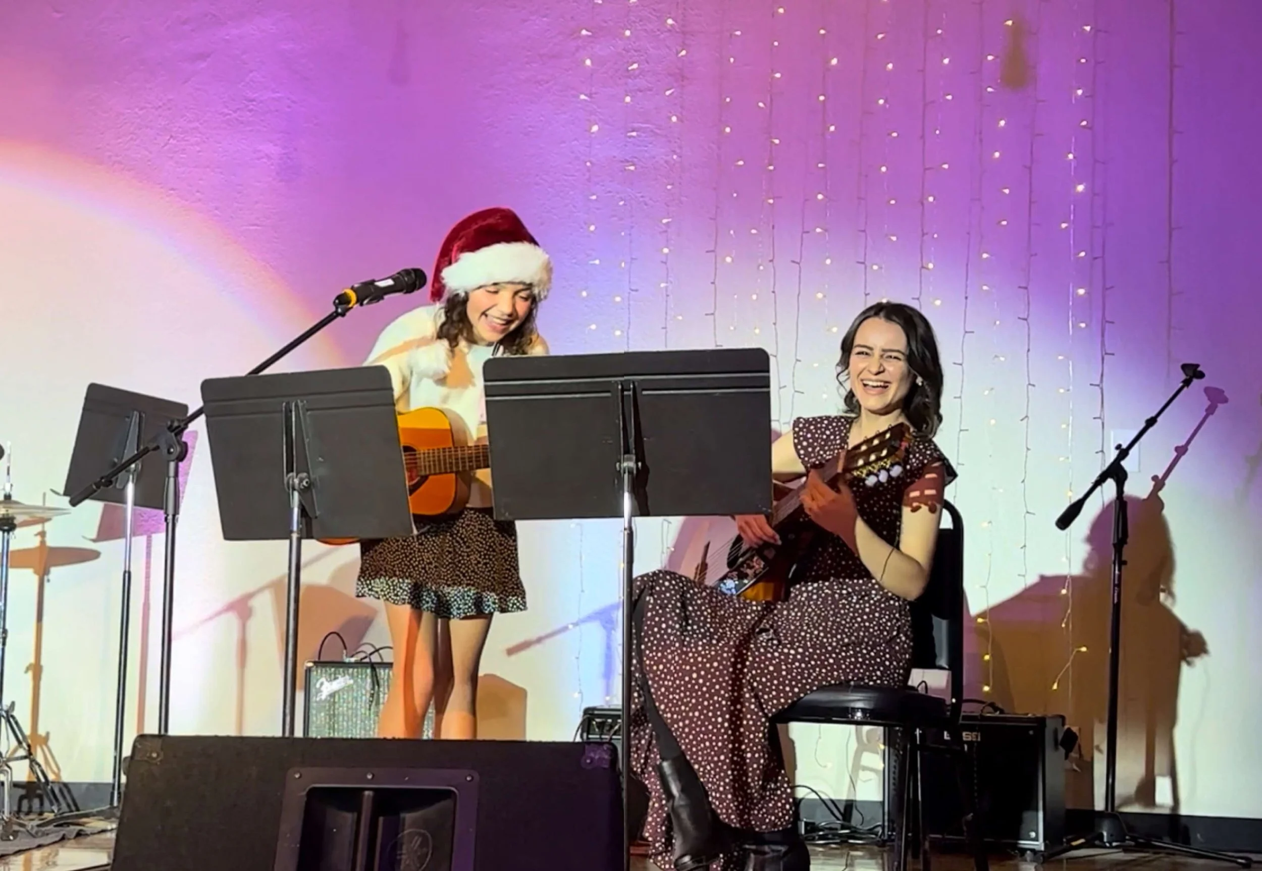 Two women performing music on stage with guitars, one wearing a Santa hat. The stage is decorated with fairy lights and microphones.
