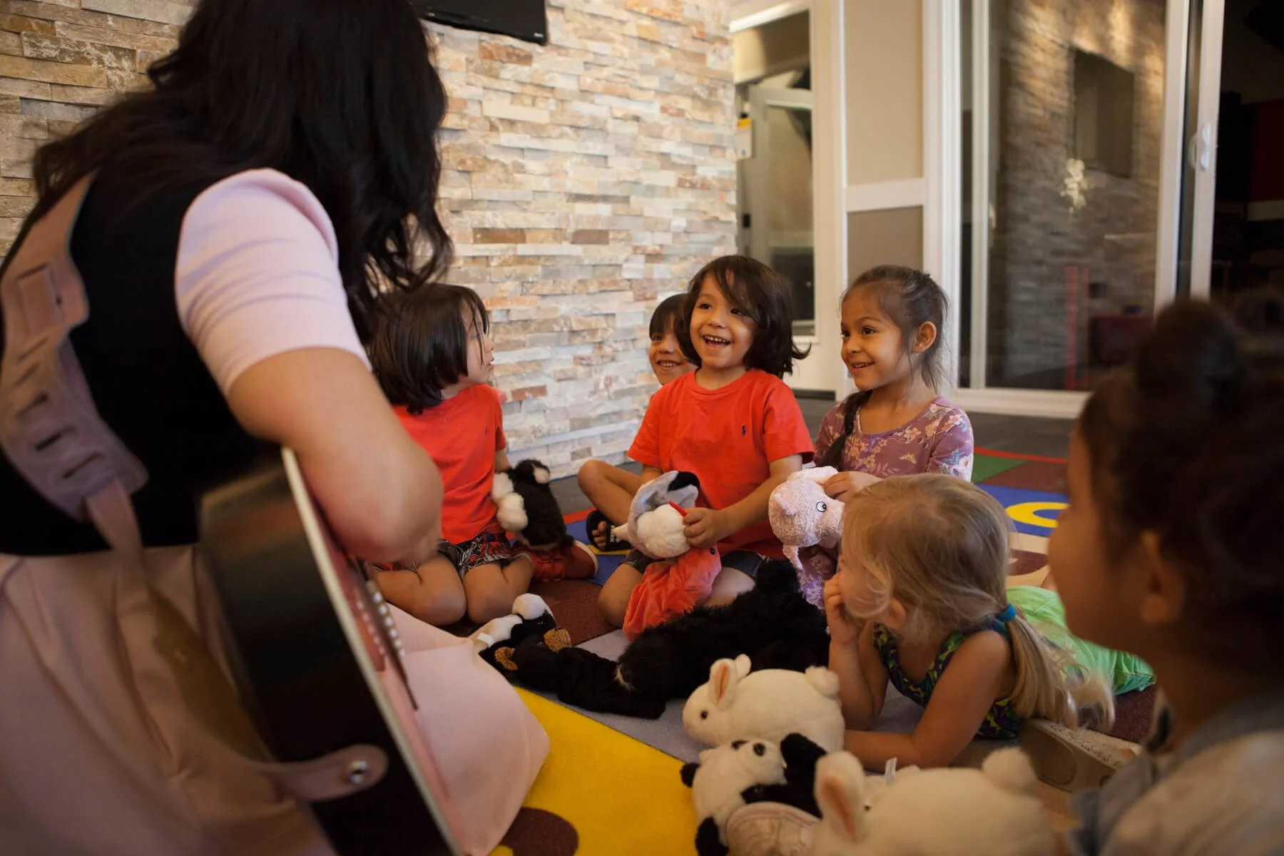 A woman playing guitar for a group of young children sitting on the floor, some holding stuffed animals, in a room with a brick wall and large windows.
