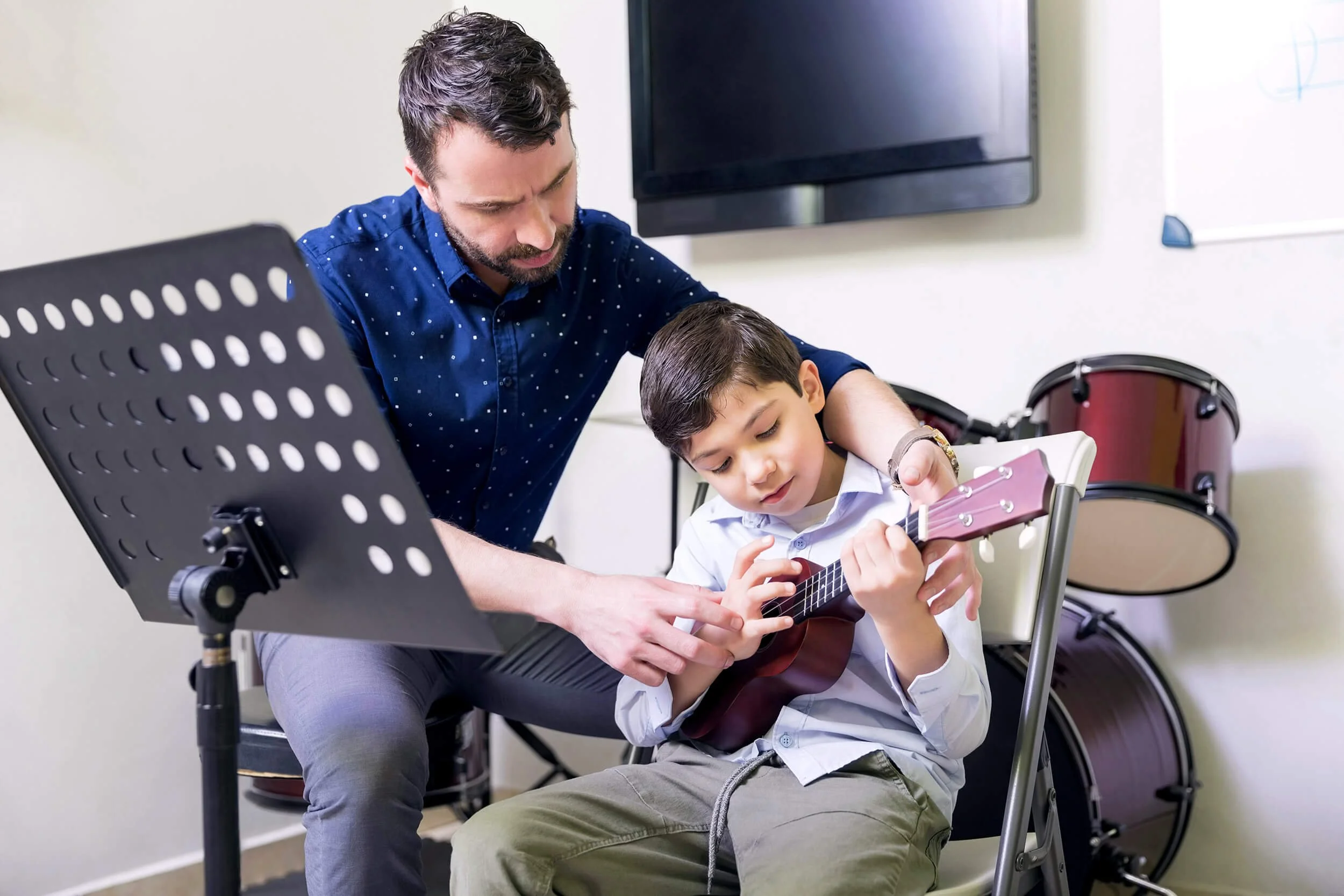 Adult man helping a young boy learn to play the ukulele in a music room with drums in the background.