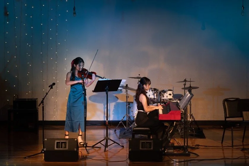 Two women performing music on a stage with a violin and piano, with drums in the background.