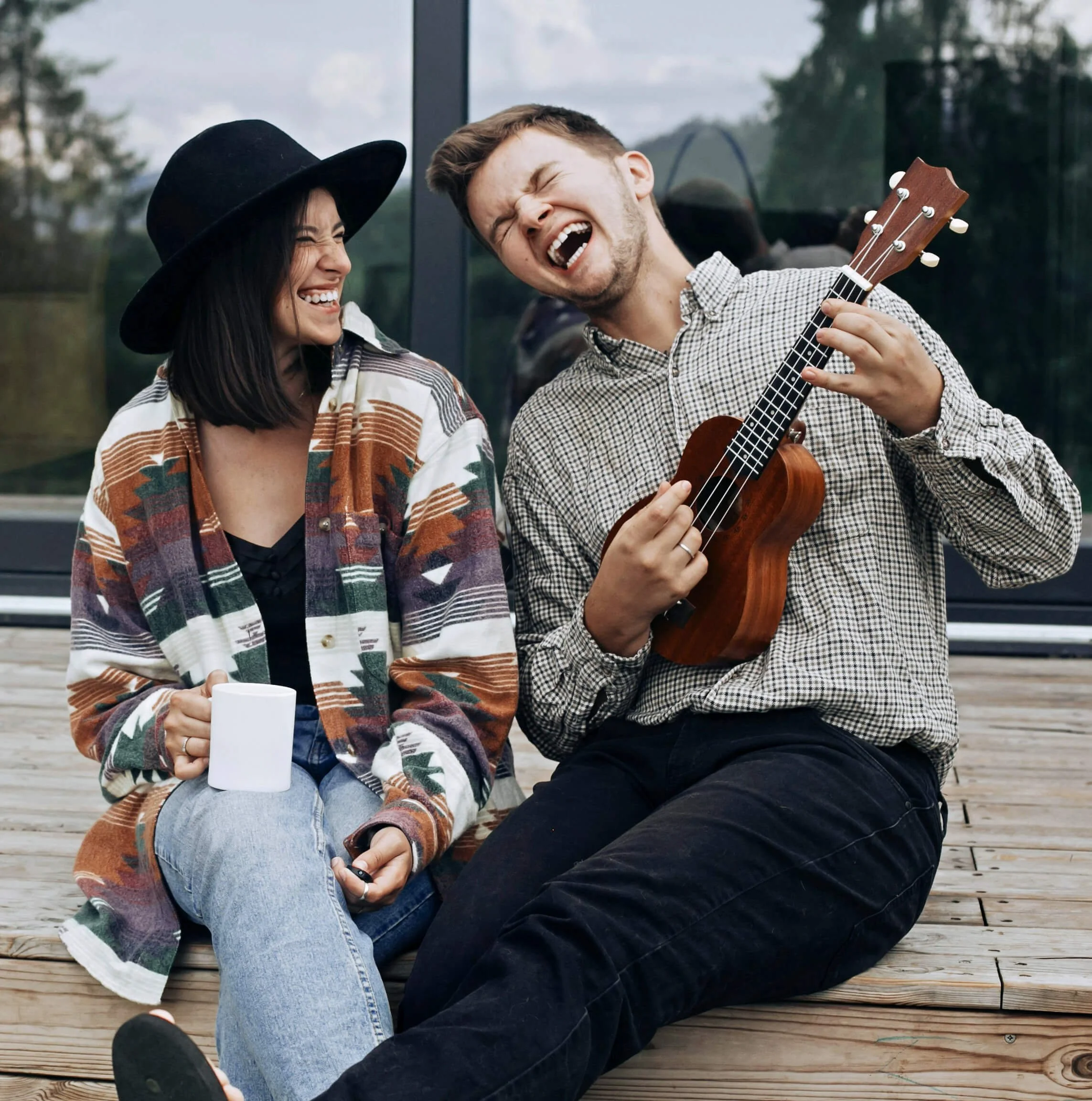 Two friends sitting on a wooden deck outdoors, laughing and enjoying music. One woman with a black hat and multicolored jacket holds a white mug, while a man with a checkered shirt plays a ukulele and sings passionately.
