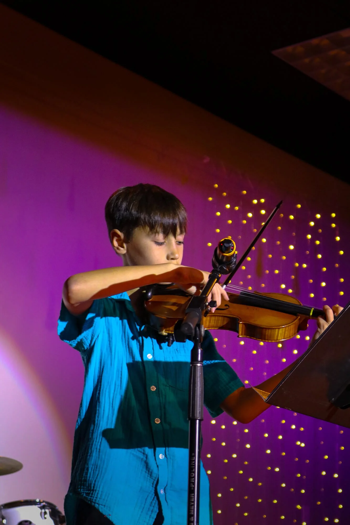 A young boy playing the violin on stage, with a music stand and purple background with yellow lights.