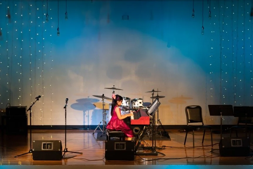 Young girl in pink dress playing a red keyboard on stage with drum set behind her, surrounded by microphones and empty chairs.