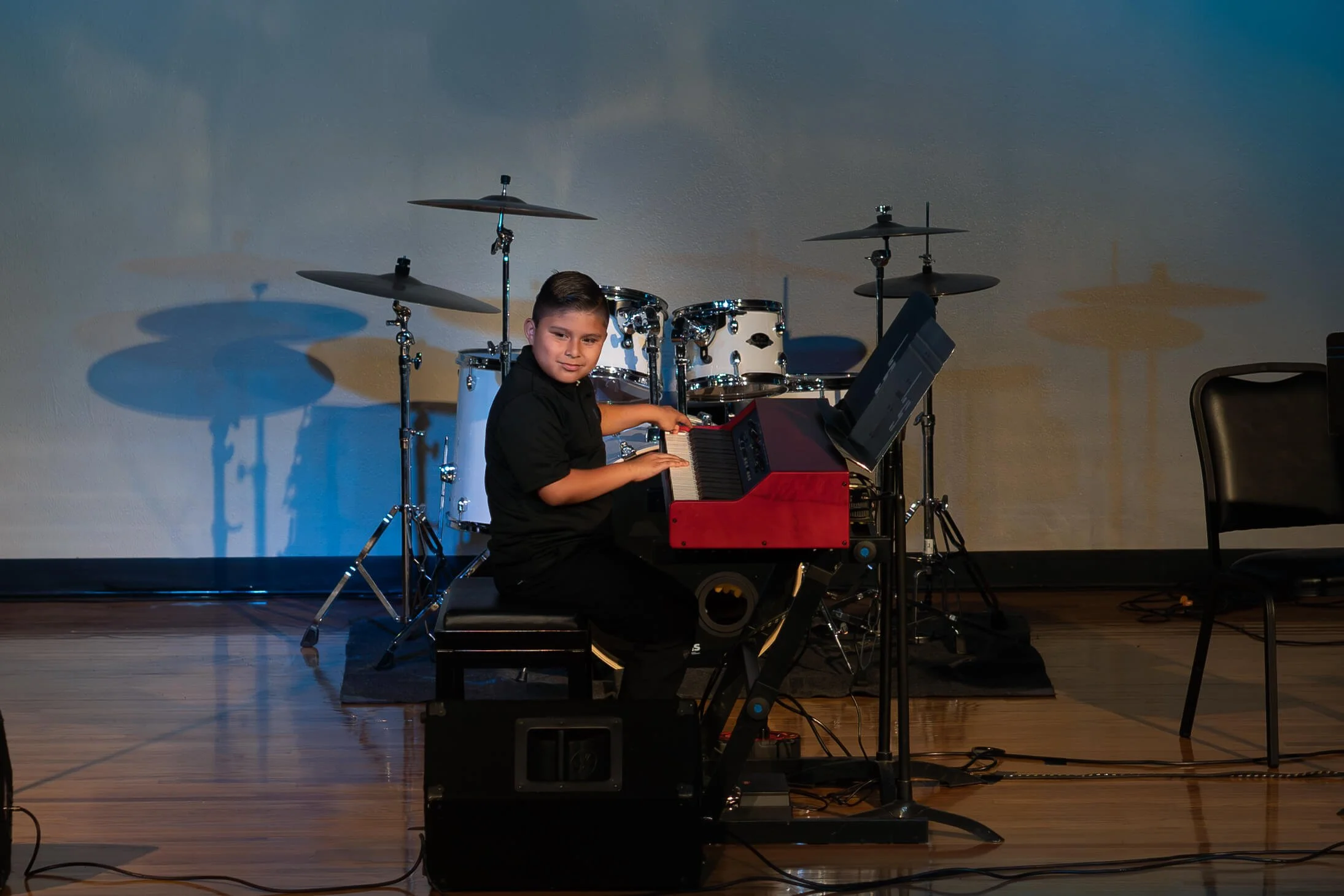 A young boy playing a red digital piano on stage with drums in the background.