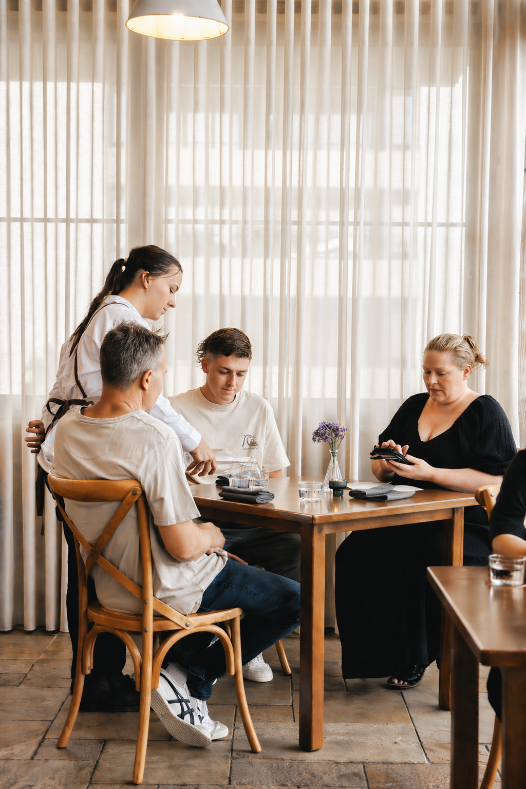 A group of five people sitting at a wooden table inside a restaurant, with a woman in a white shirt gesturing towards an older man, a young man, a woman in black looking at a phone, and another person partially visible on the right. There is a vase with purple flowers on the table, and the background features sheer curtains and a ceiling lamp.