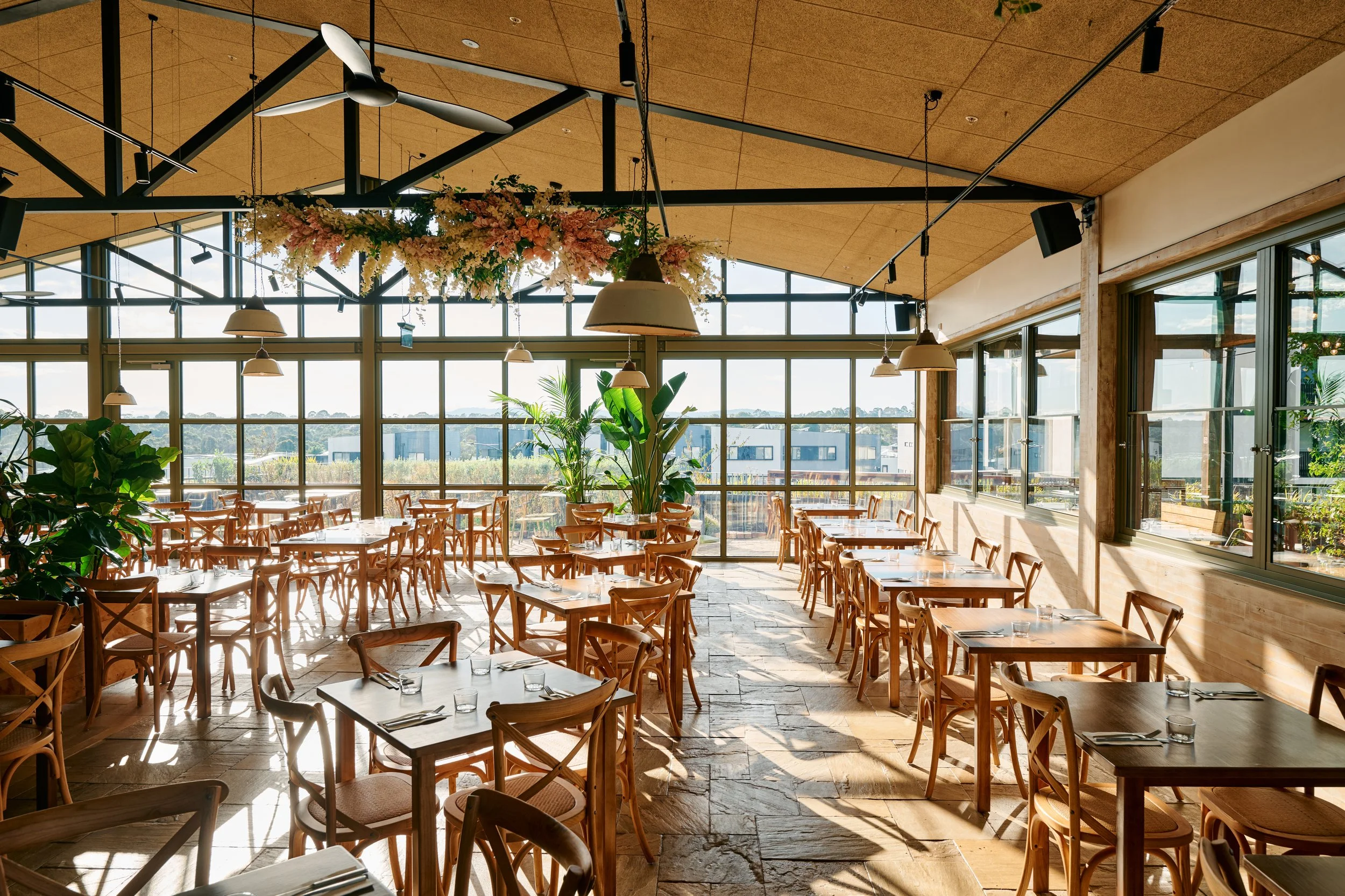Interior of a modern restaurant with large windows, wooden tables and chairs, hanging lamps, and decorative plants, with sunlight streaming in.