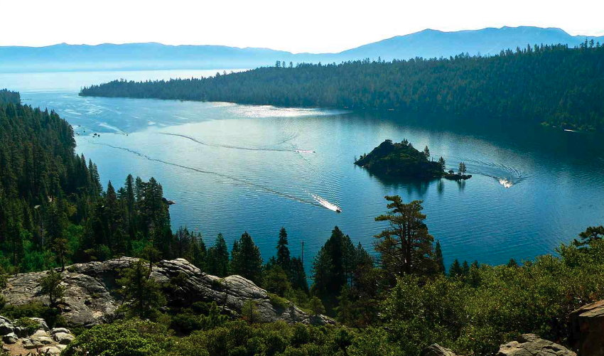 A panoramic view of South Lake Tahoe surrounded by dense green pine forests, with boats creating wakes on the water and distant mountains in the background.