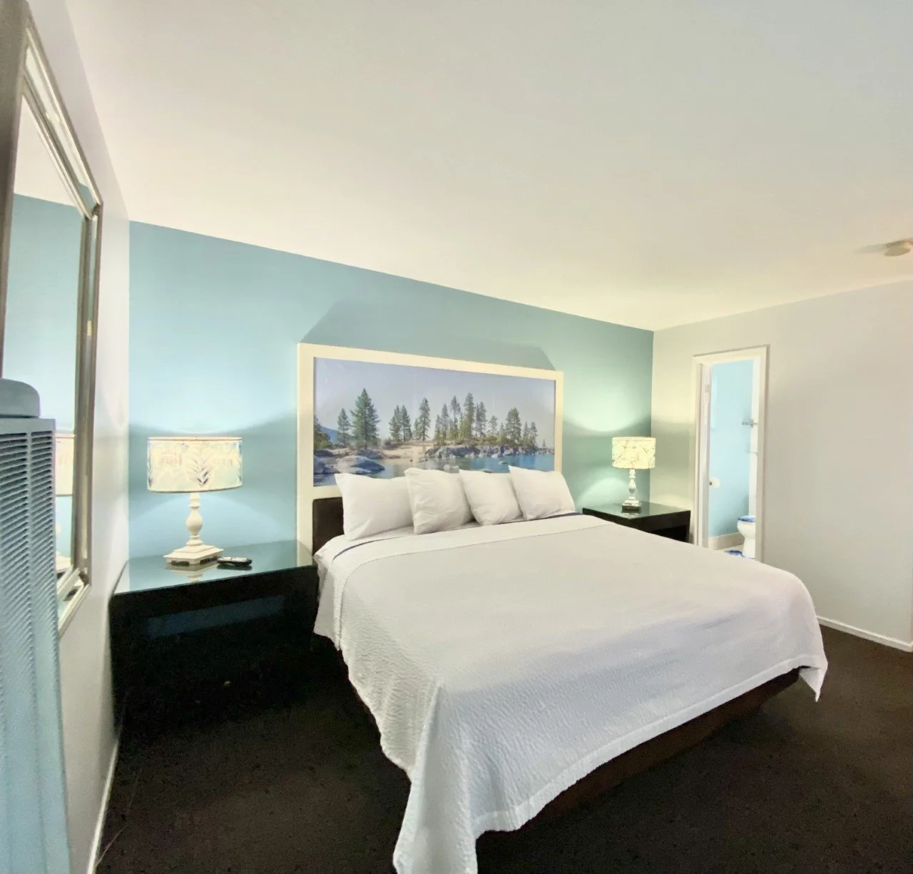Hotel room with a large King bed, white bedding, pillow arrangement, artwork of trees and lake above the headboard, two bedside lamps, and a bathroom.