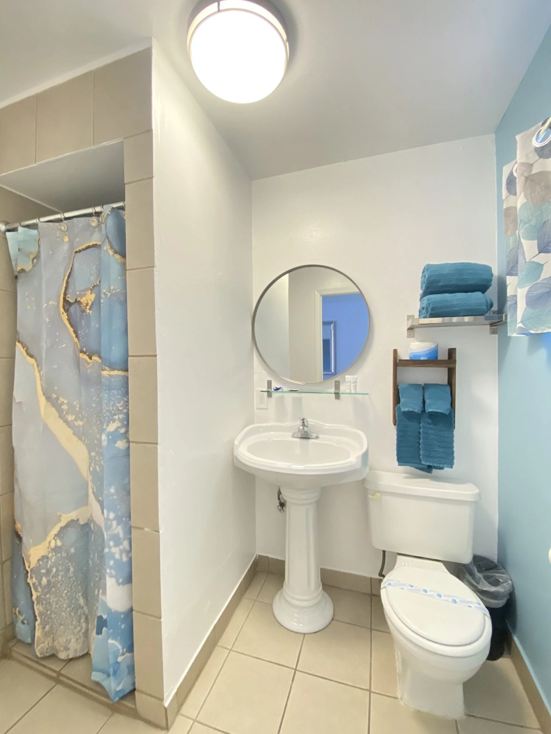 Bathroom with a pedestal sink, oval mirror, toilet, and a shower with a blue and gold patterned curtain. Blue towels are on a wall-mounted shelf and towel rack. There is a ceiling light and light blue walls.