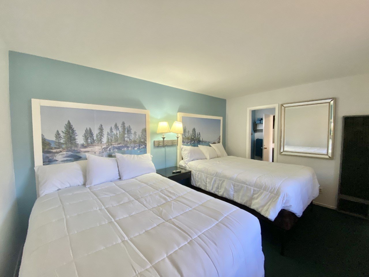 A hotel room with two Queen beds, white bedding, lake and trees artwork on the headboards, a wall-mounted lamp between the beds, a mirror on the wall, and a doorway leading to another room at Bluebird Day Inn in South Lake Tahoe CA