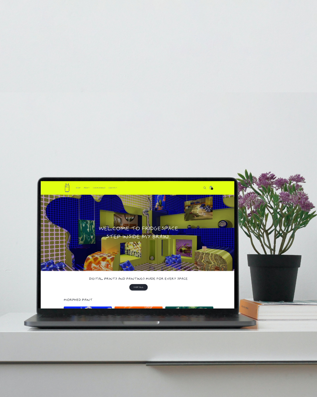 Laptop displaying a vibrant art-themed website with digital prints and paintings, placed on a white desk next to a potted green plant and a closed book.