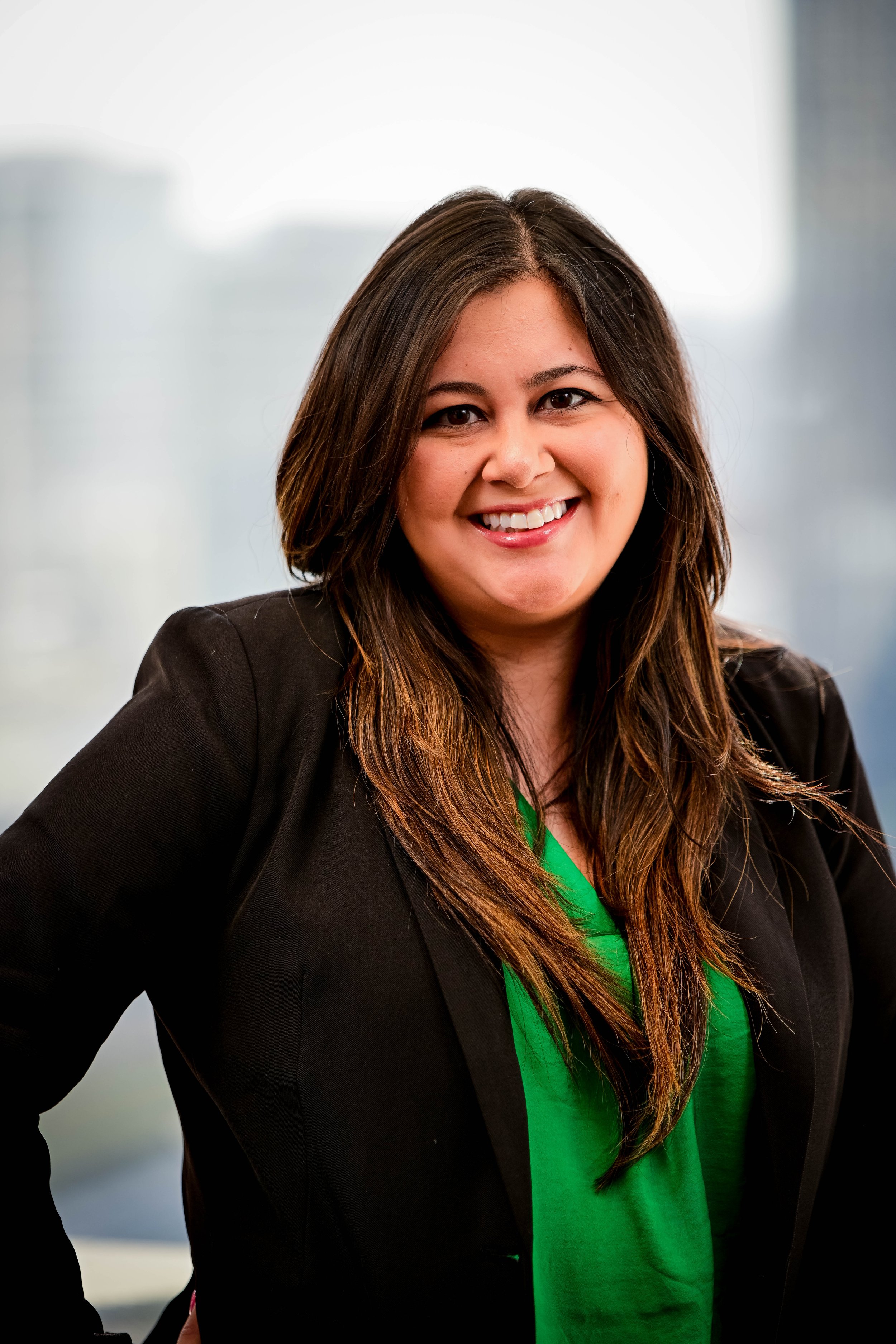 A woman with long brown hair, smiling, wearing a black blazer over a green shirt, standing indoors with a blurred cityscape background.