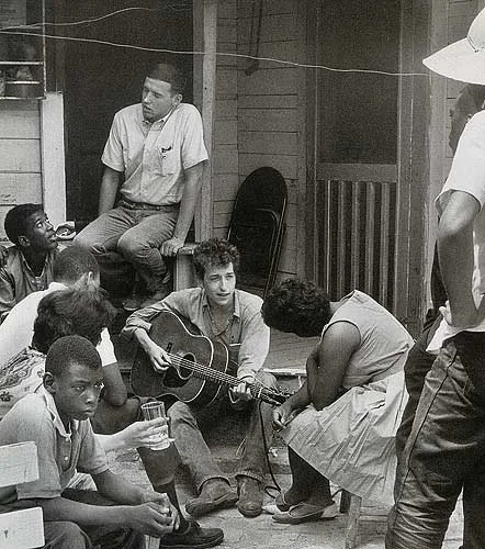 A black and white photograph of a young Bob Dylan playing the acoustic guitar on a front porch