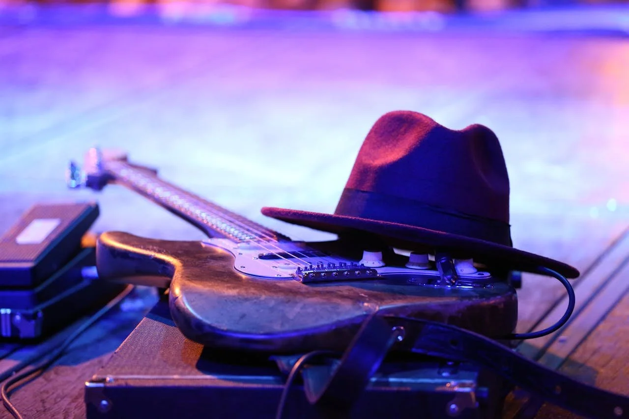 A photograph of an electric guitar laid on a stage, a fedora resting on top