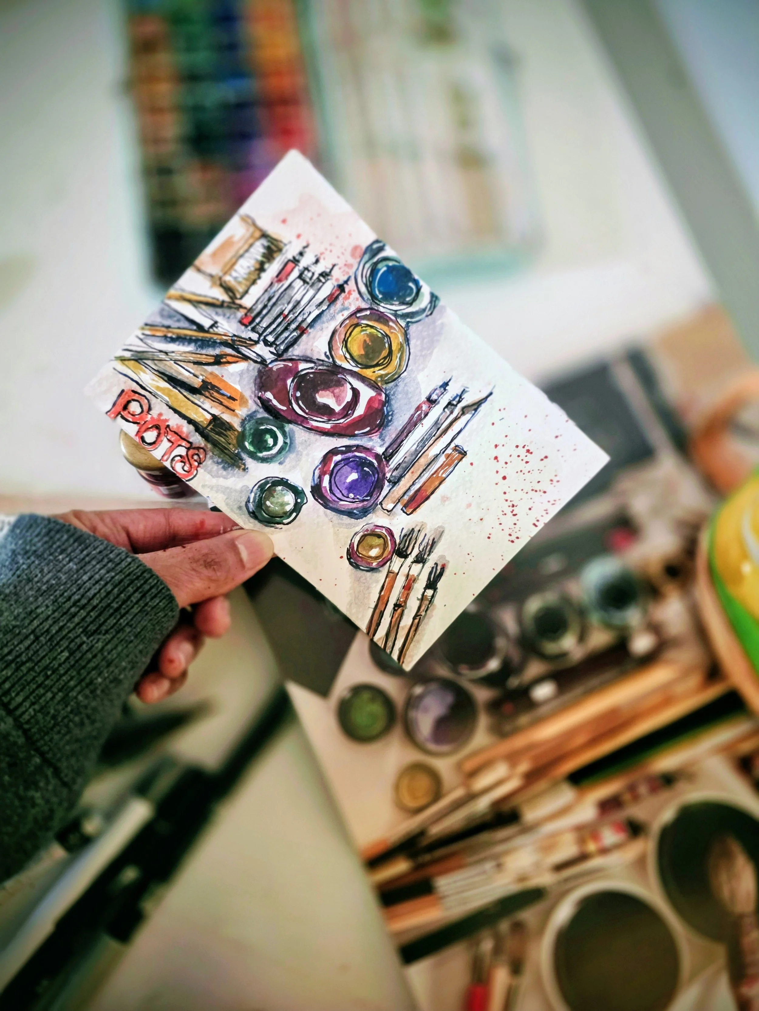 A hand holding a small piece of paper with a colorful watercolor painting of art supplies, including paint pots, brushes, and a palette, with the word 'arts' written on it. In the background are containers of watercolor paints.
