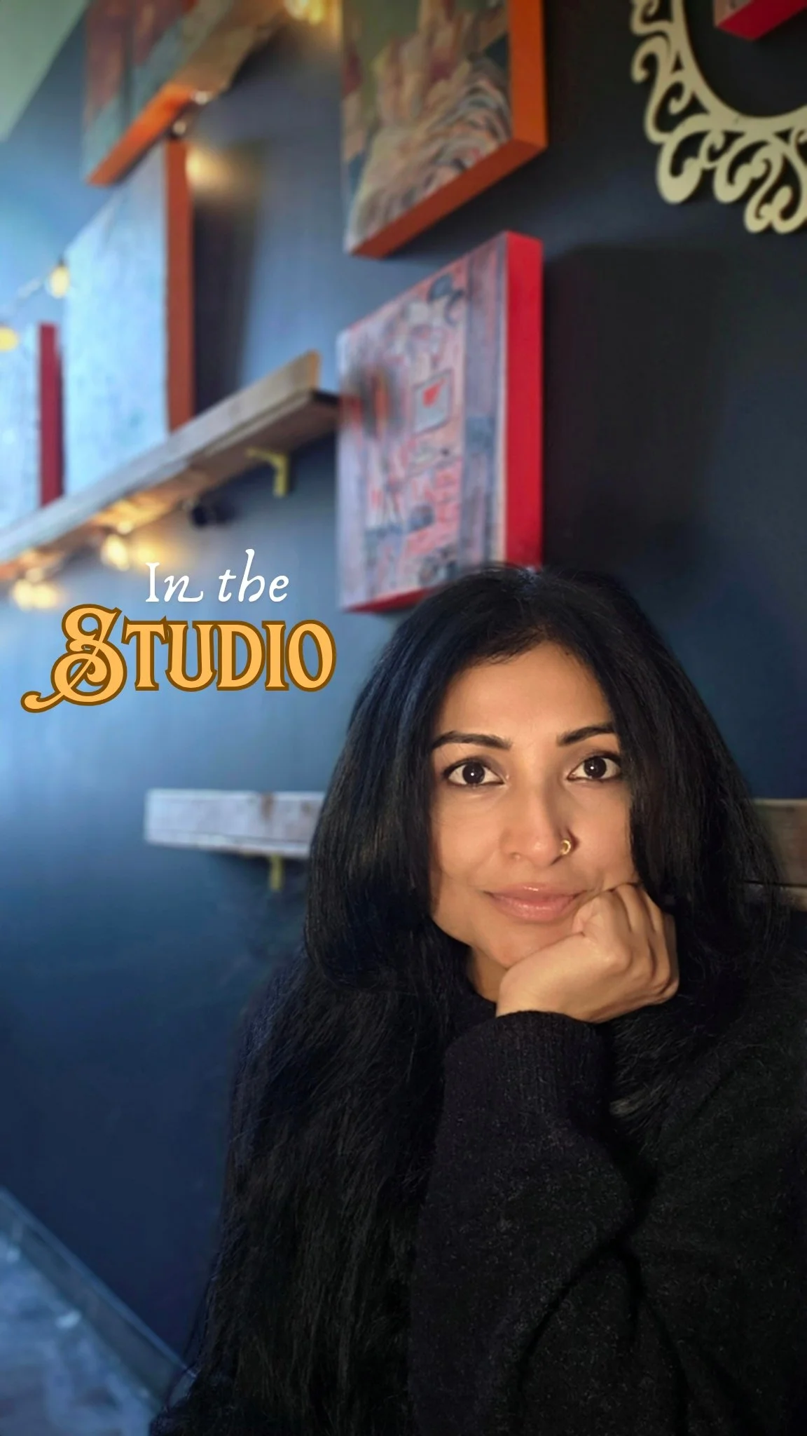 A woman with long black hair and a nose ring sitting in a studio, with a dark wall behind her decorated with colorful paintings and a decorative wall hanging. The image has text saying "In the STUDIO".