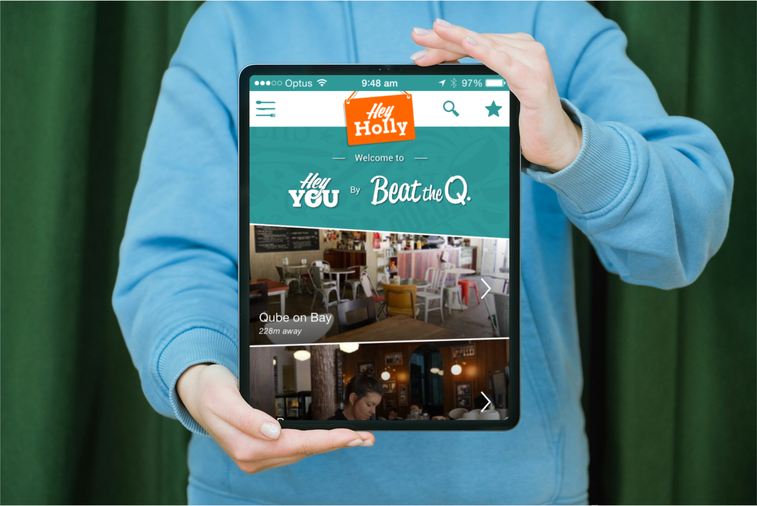 Person in a blue hoodie holding a tablet displaying Hey You app with pictures of cafes and text.