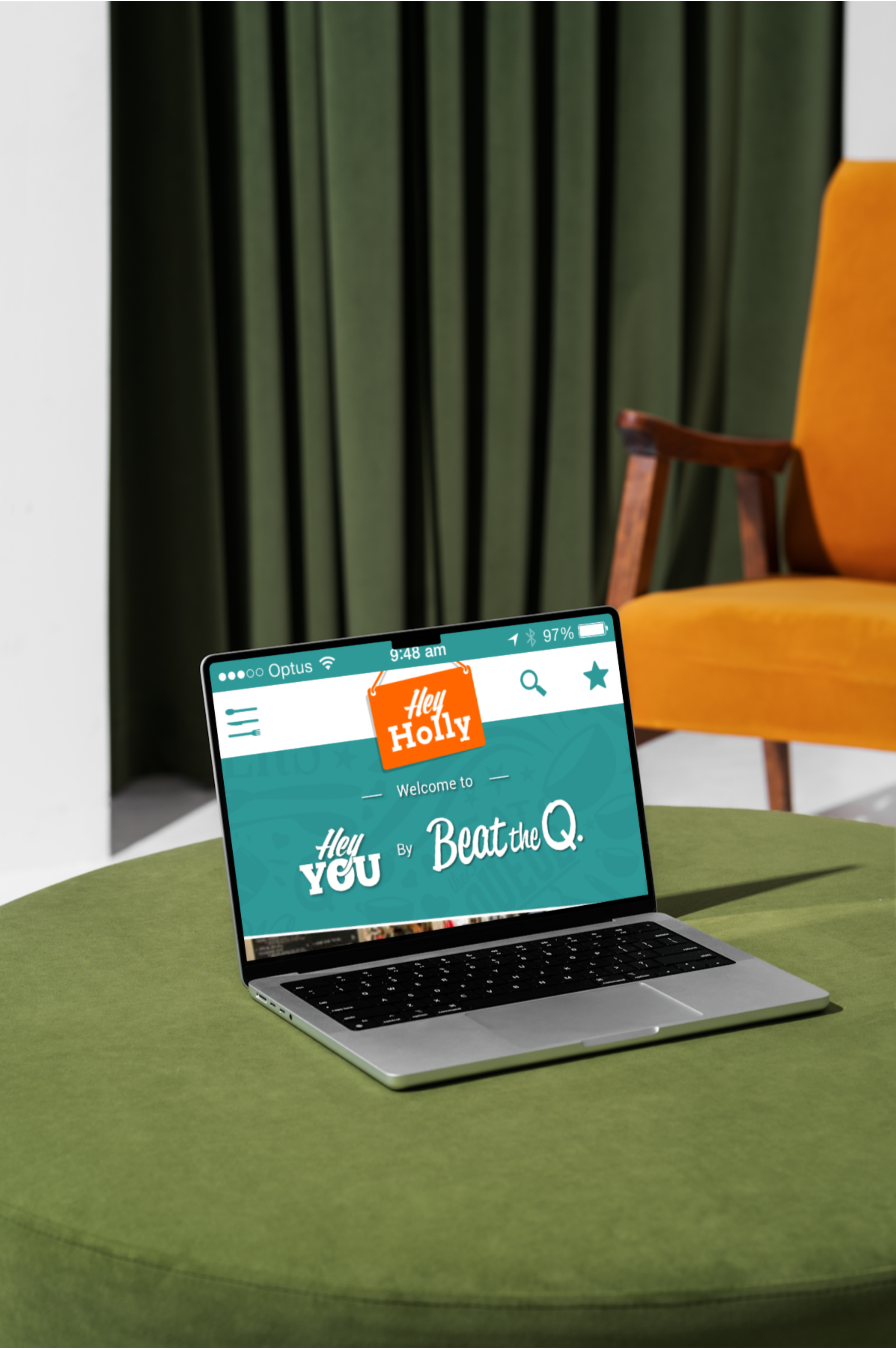 A laptop on a green table displaying the Hey You website with a welcoming message, surrounded by a yellow chair and a green curtain in the background.