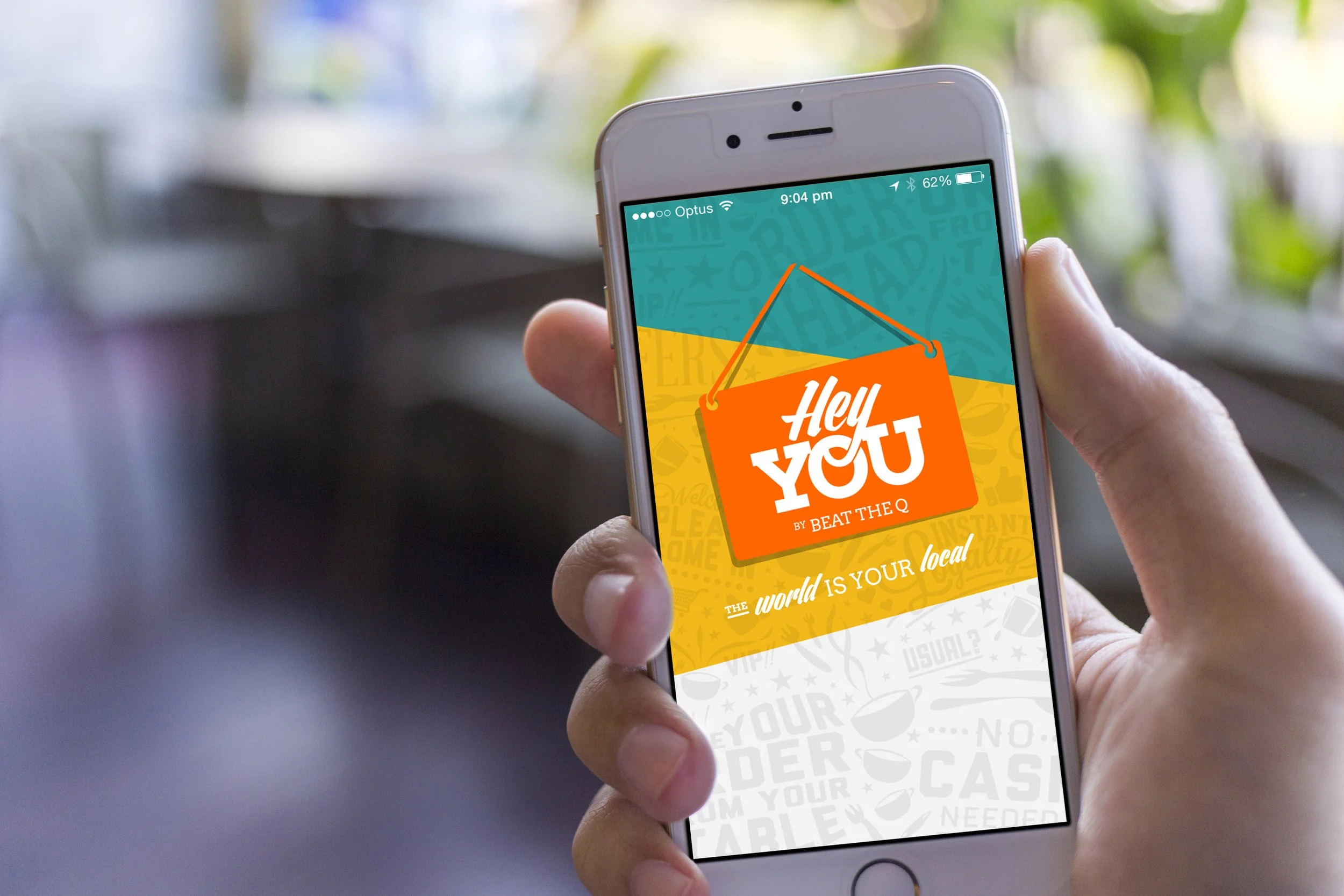 A person holding a smartphone displaying a colorful app with the text 'Hey You by Beat The Q' and the slogan 'The world is your local'.