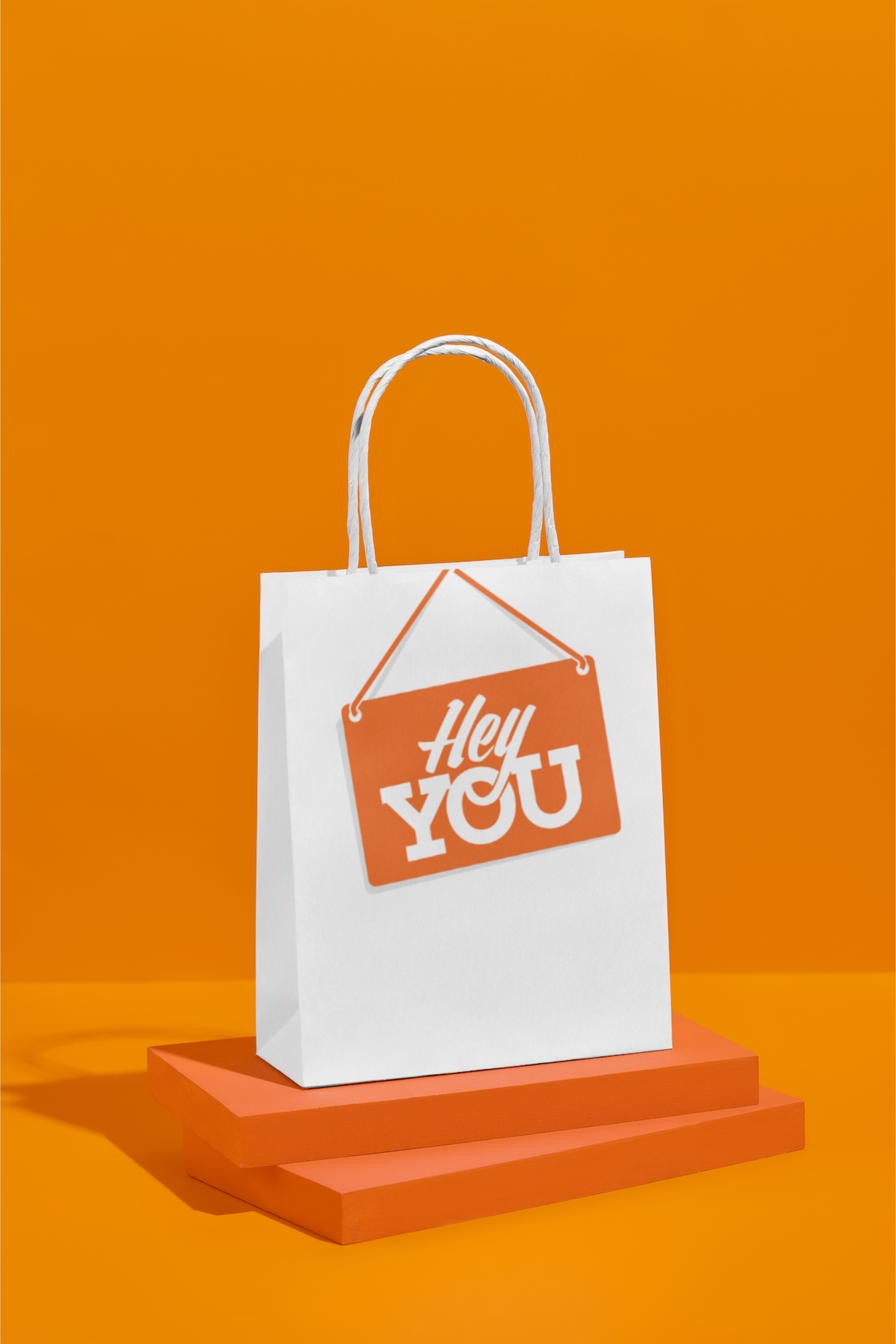 A white shopping bag with orange handles and an orange sign that reads 'Hey You' hangs on an orange wall, placed on orange pedestals.