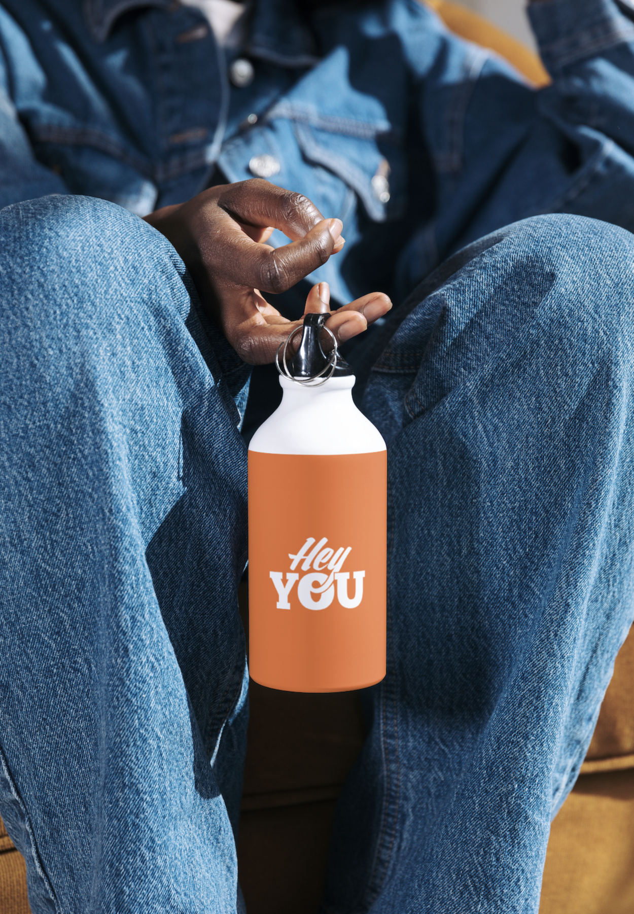 Person sitting with legs crossed holding a spray bottle with a label that says 'Hey You' in a warmly lit room.