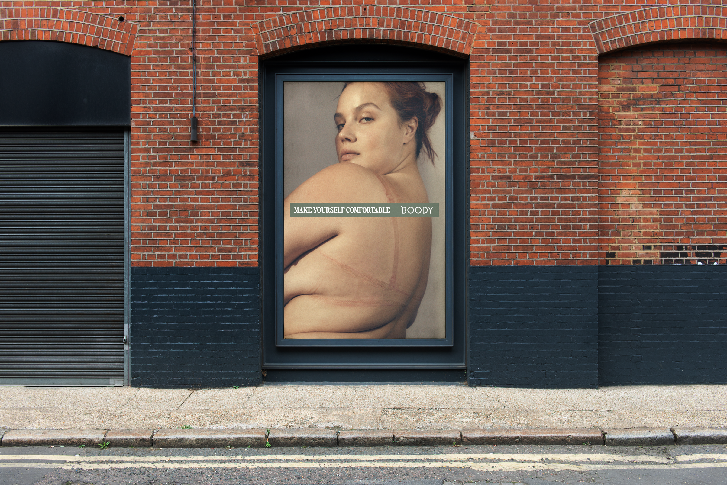 A large poster on a brick wall featuring a woman with red hair looking at the camera, with the text 'Make Yourself Comfortable' and 'Boody' across her shoulder and arm.