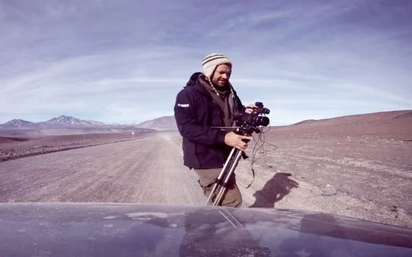 A man standing outdoors in a barren, mountainous landscape holding a camera mounted on a tripod.