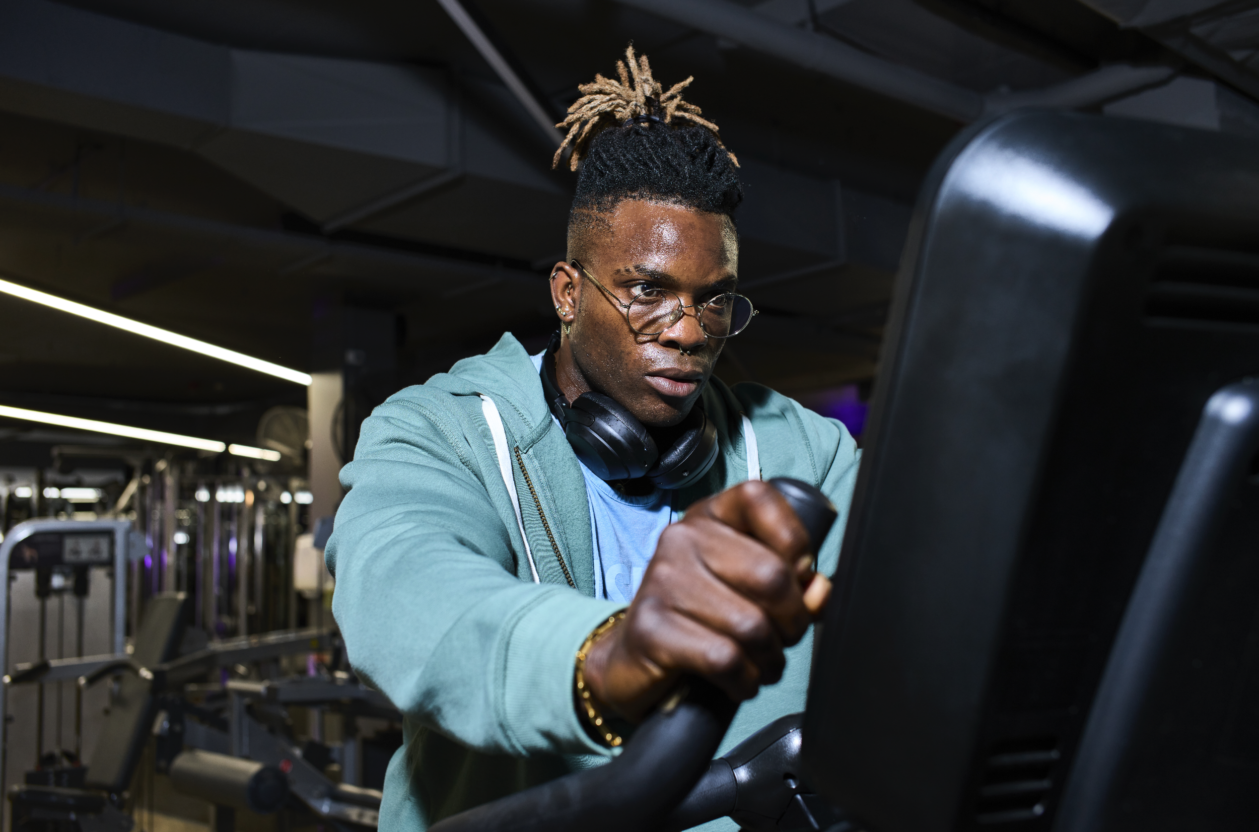 A man with glasses and braided hair at a gym, wearing headphones around his neck, working out on a treadmill.