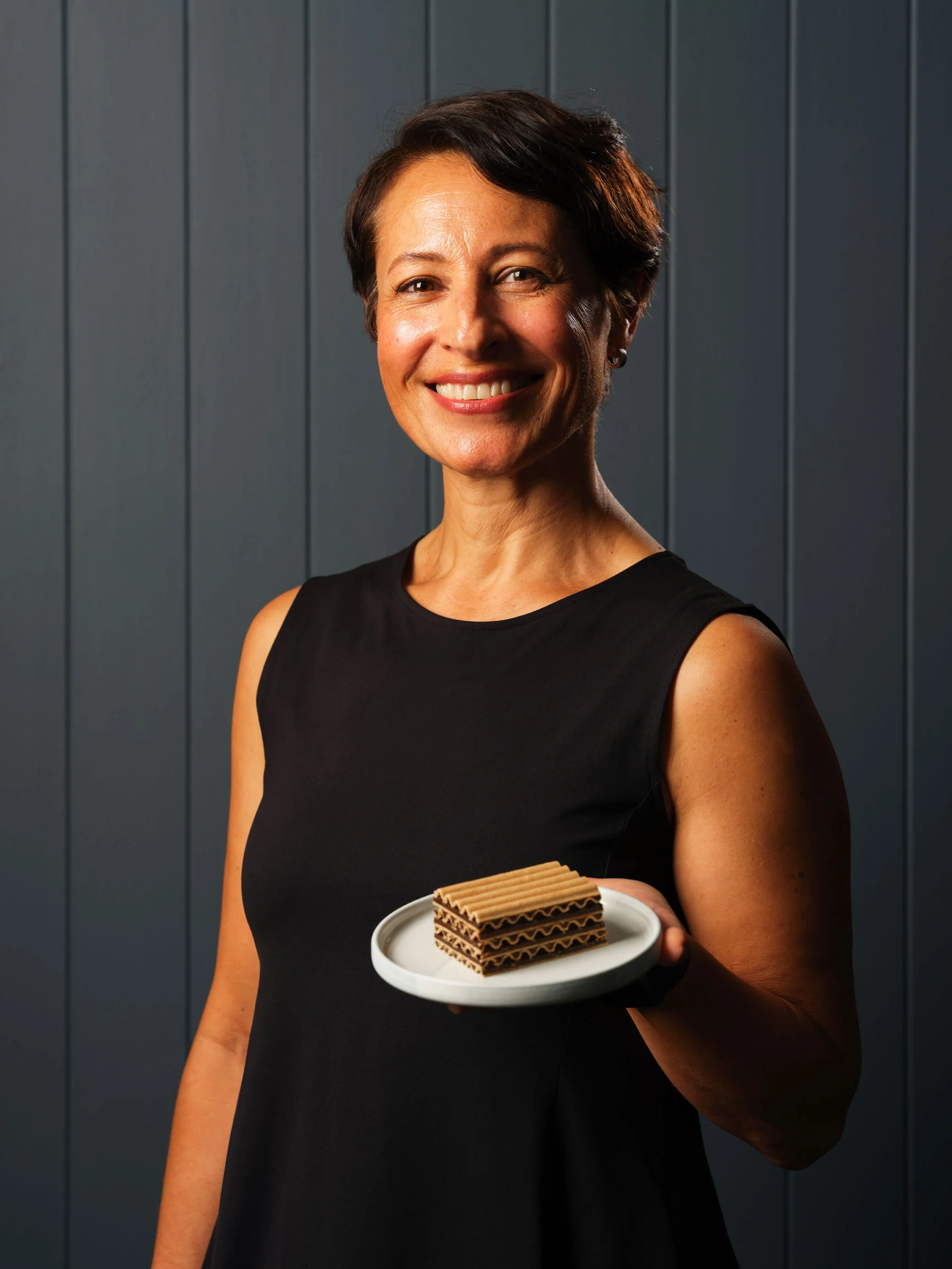 A woman with short dark hair smiling, wearing a black sleeveless dress, holding a small white plate with layered chocolate wafers, standing in front of a dark gray paneled wall.