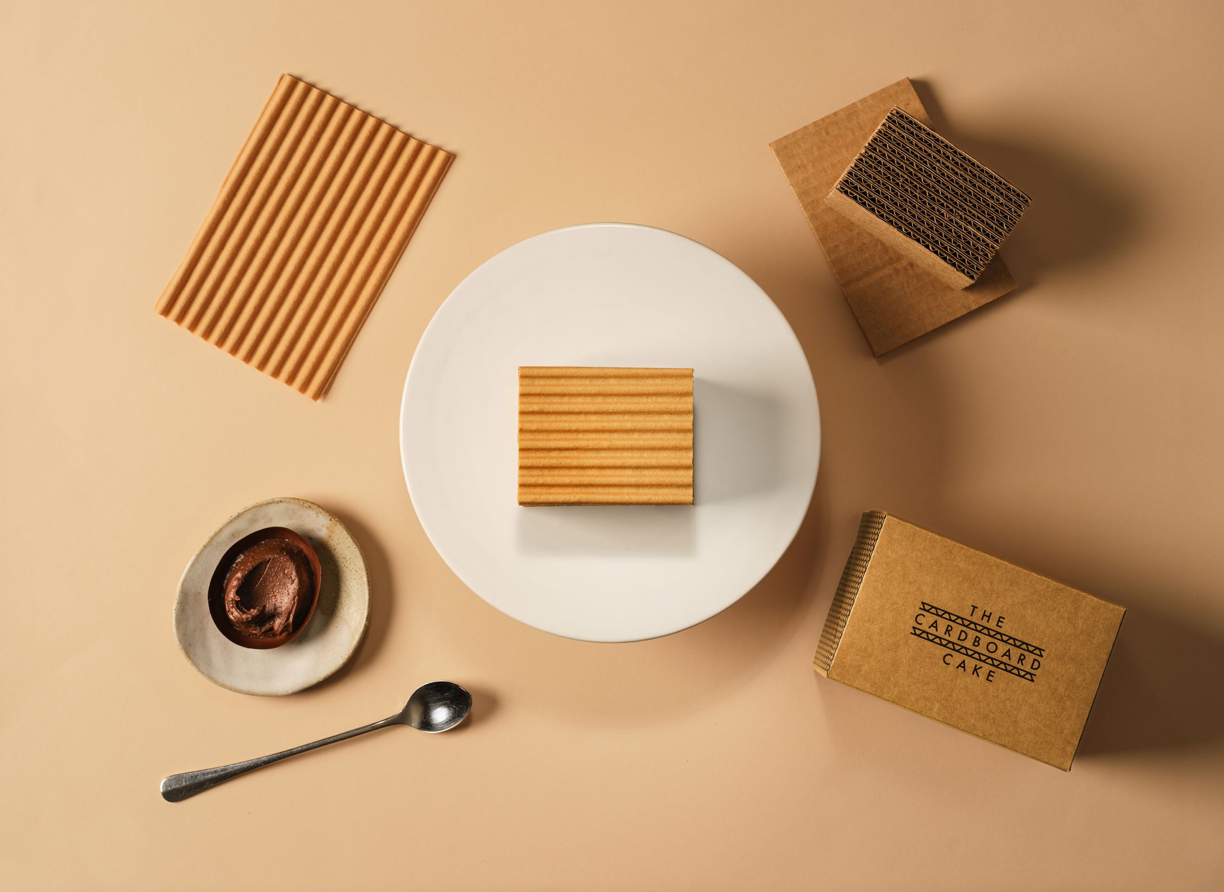 A wooden layered cake square on a white plate, with additional wooden cake slices and packaging boxes around, and a spoon with ice cream in a small dish on a beige background.