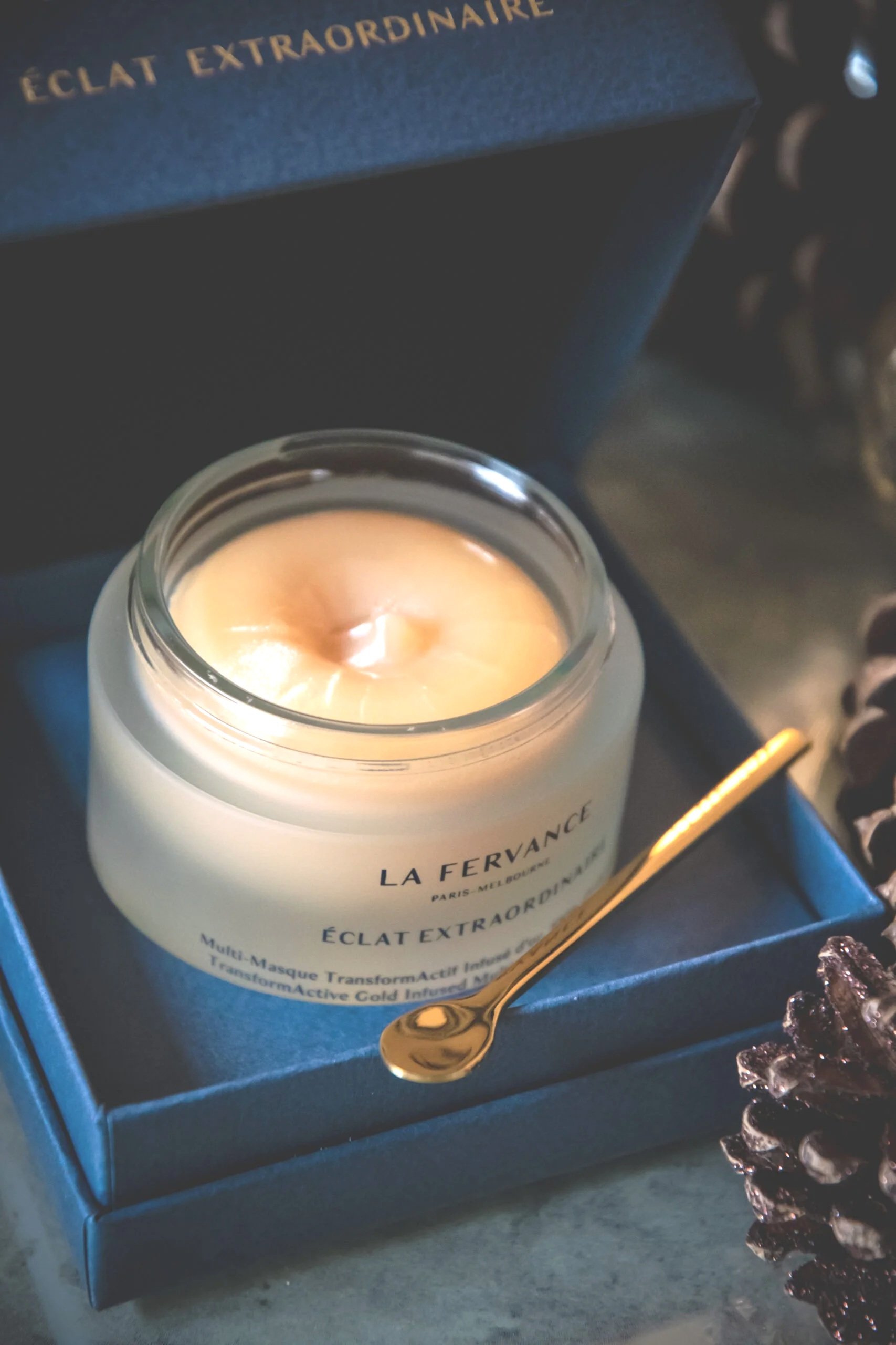 A lit candle in a glass jar with gold writing, placed in a blue box with a gold spoon nearby. The candle label reads 'LA FERVANCE.' Part of the blue box has the words 'ÉCLAT EXTRAORDINAIRE' in gold.