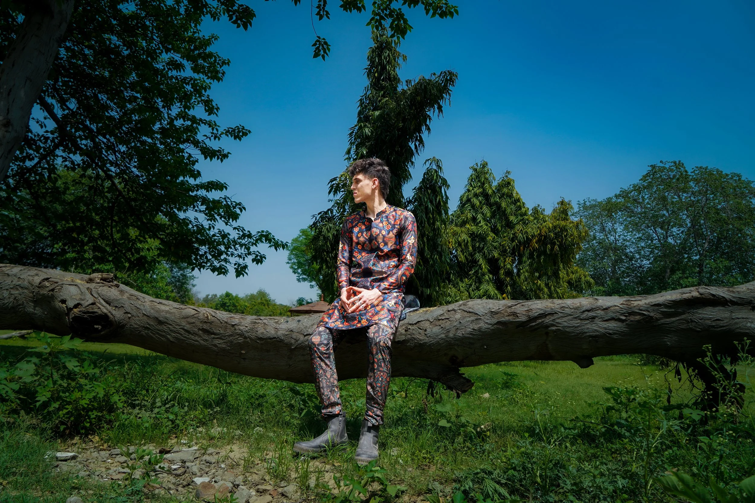 A person in a patterned kurta sitting on a fallen tree trunk outdoors, surrounded by green trees and a blue sky.