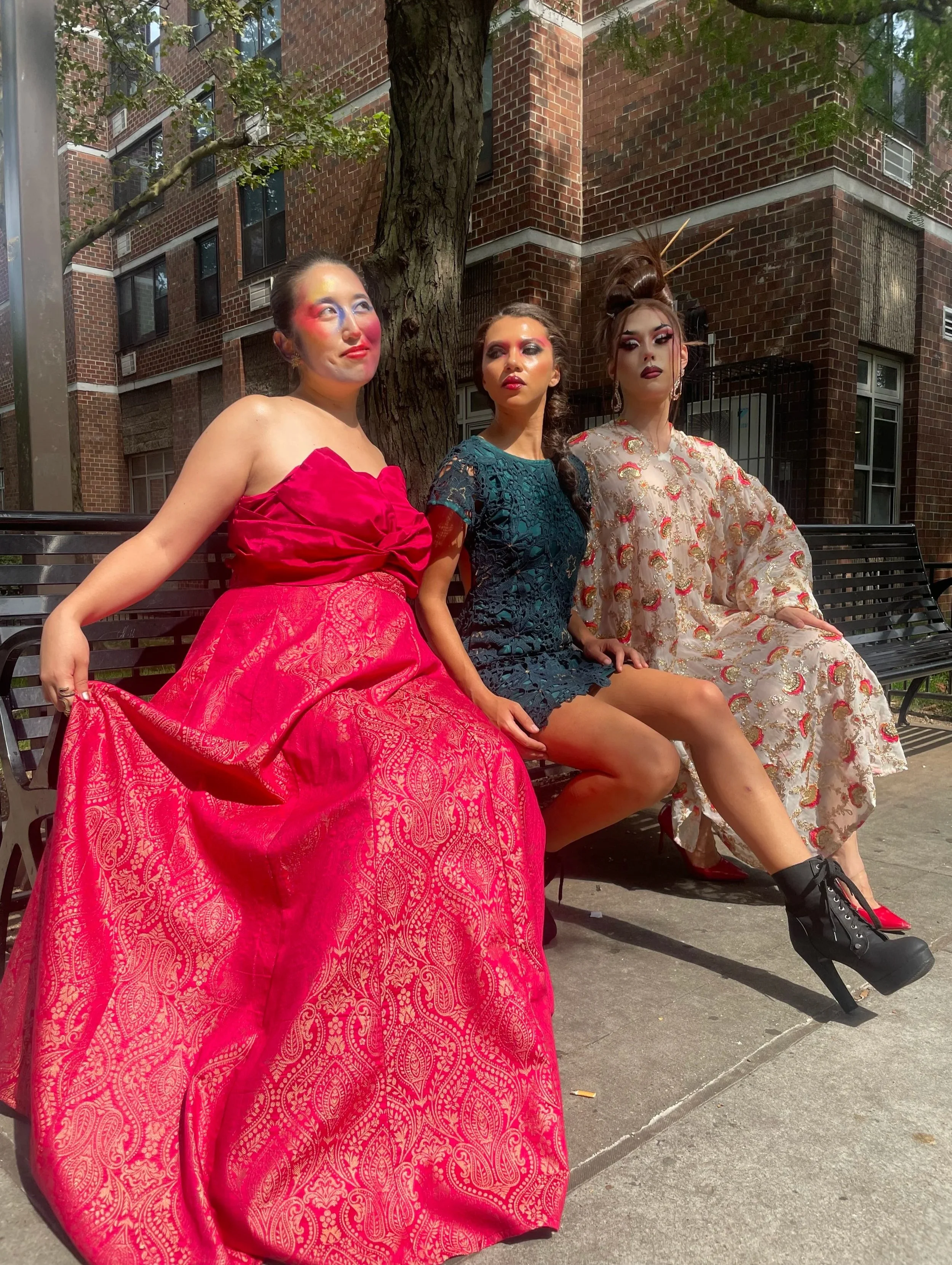 Three women sitting on a park bench in front of a brick building and tree, dressed in avant-garde fashion with colorful makeup and hairstyles.