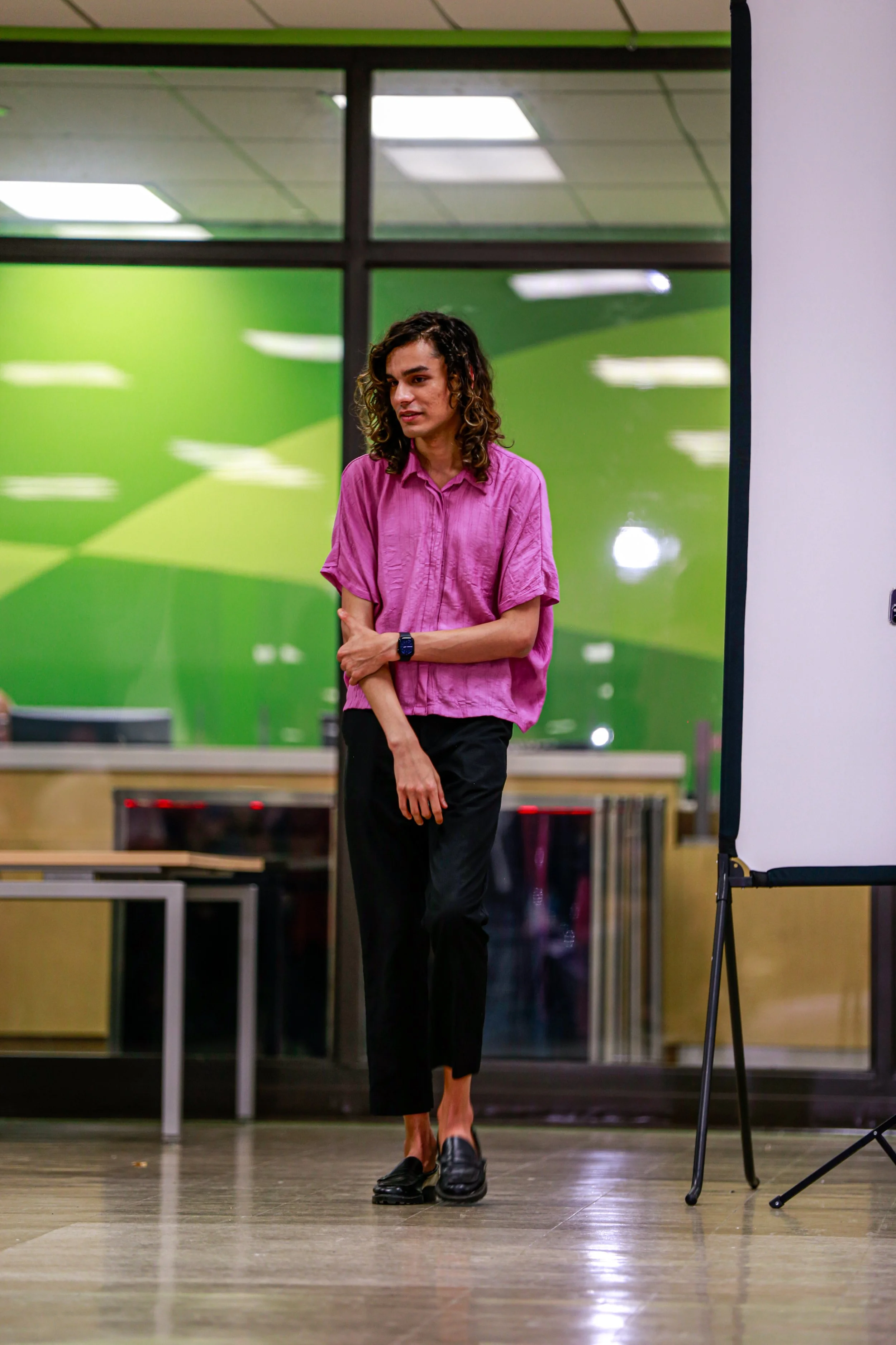 A person standing in front of a presentation screen with a wood floor, wearing a pink shirt, black pants, and black shoes, in a room with reflective glass walls and green-colored background.