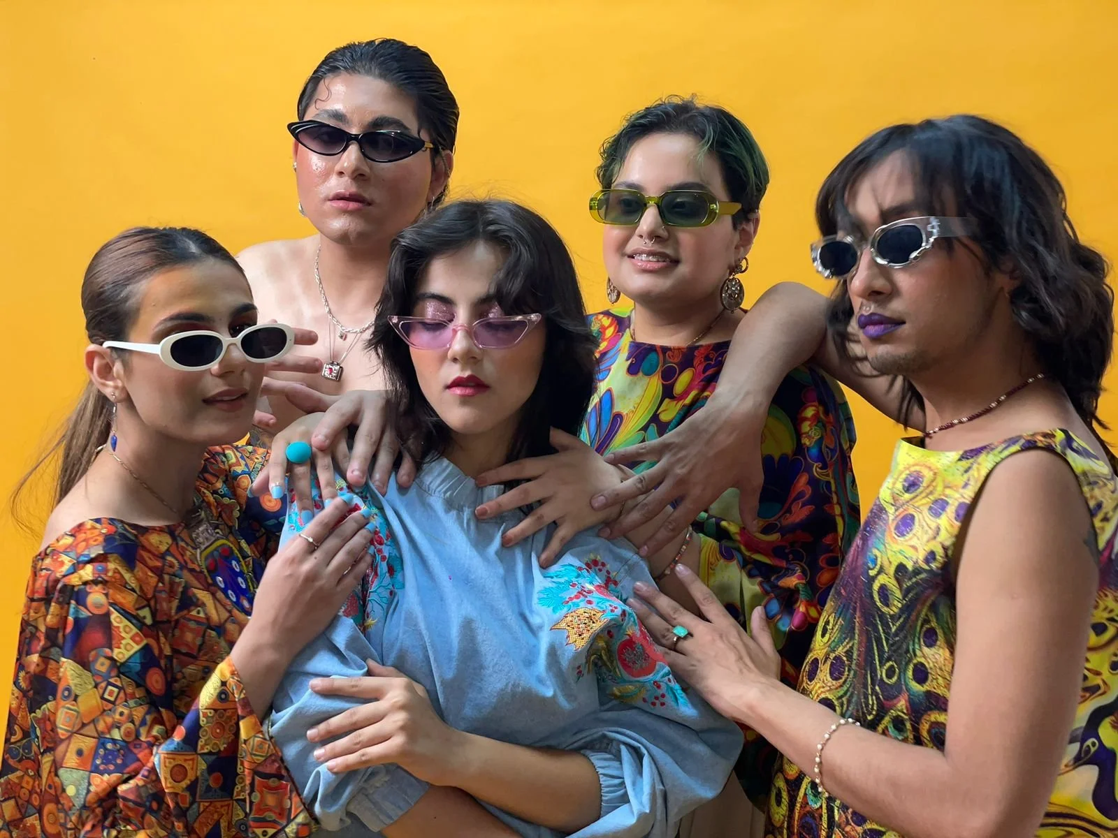 A group of five fashion models dressed in colorful, patterned clothing, wearing sunglasses, and playfully posing together against a yellow background.