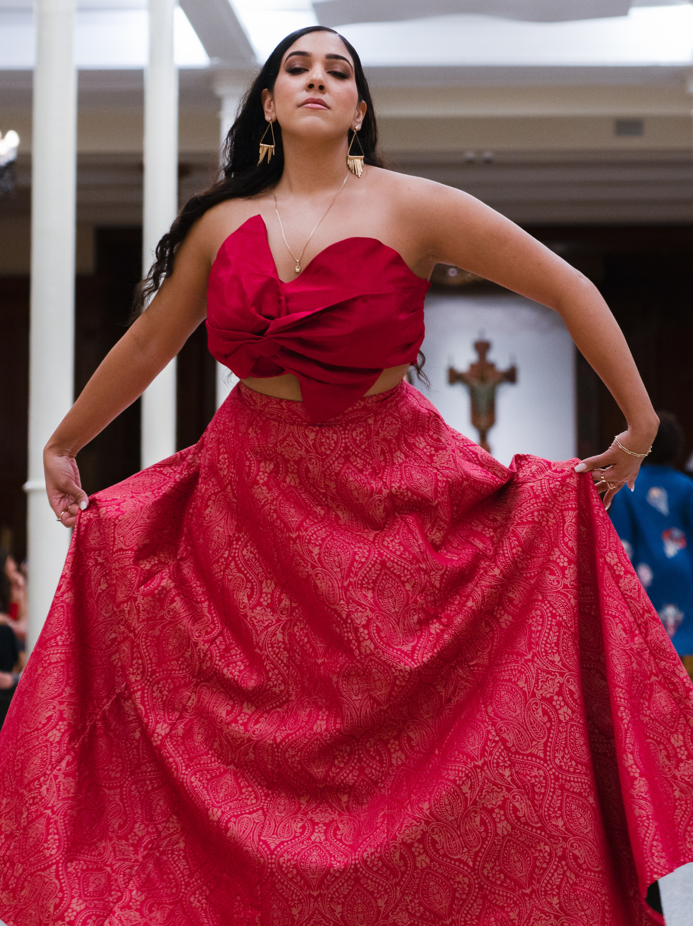 A woman wearing a red strapless dress with a large bow on the top and an intricate patterned skirt, holding out the sides of her gown, in an indoor setting with vertical columns.
