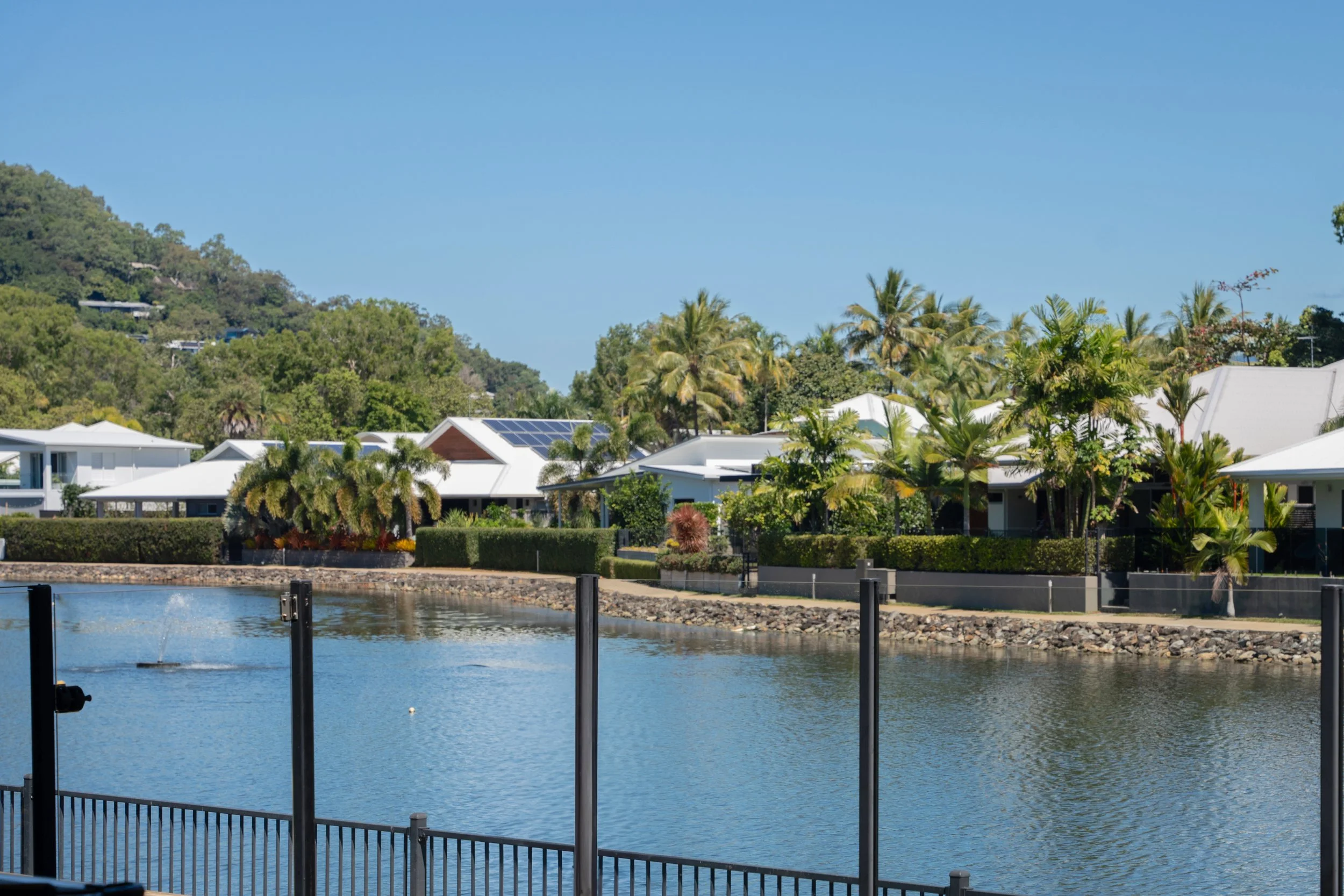 Calm water with a railing in the foreground, modern houses with solar panels, lush greenery, palm trees, and a hillside in the background under a clear blue sky.