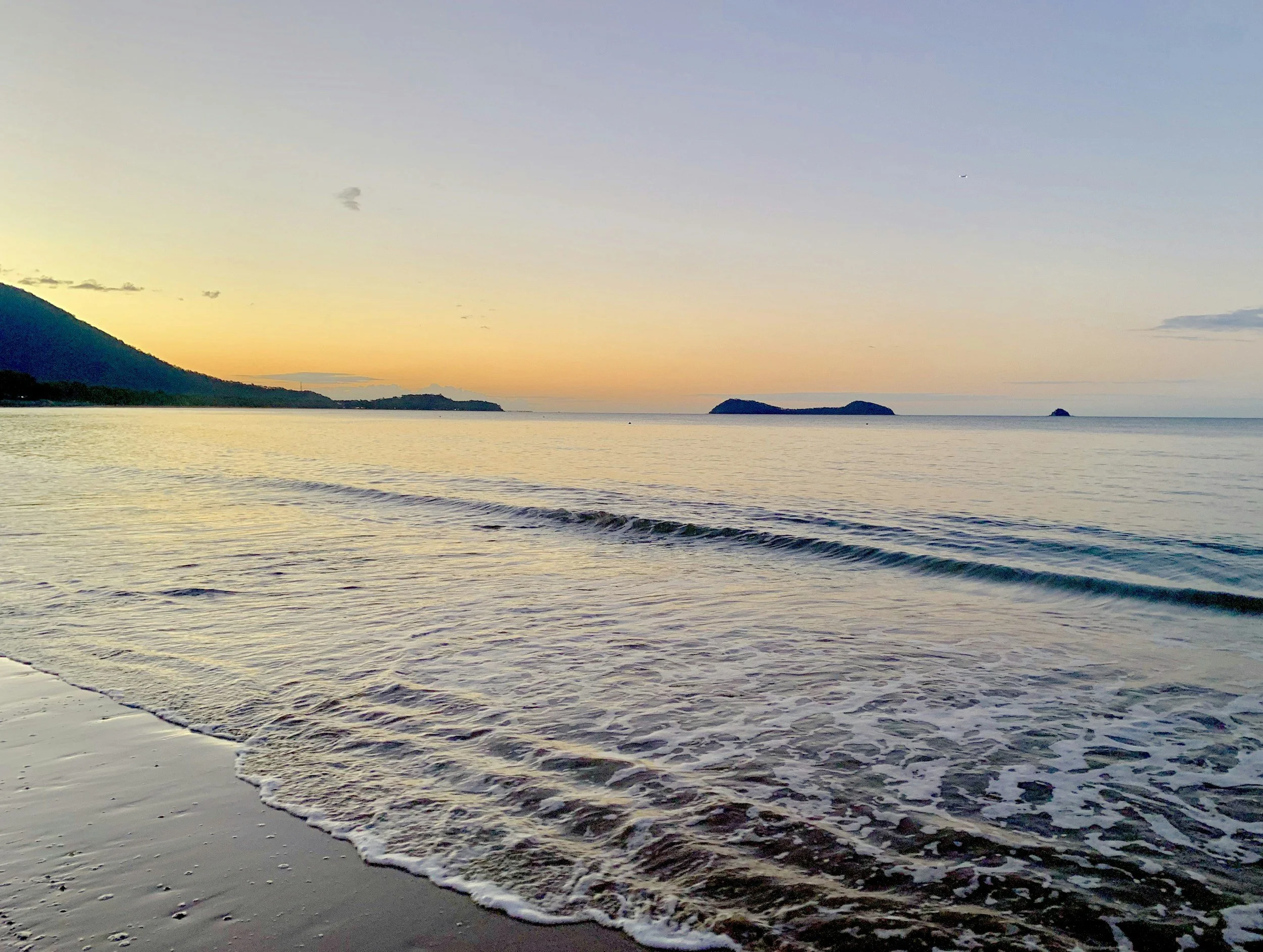 A calm ocean shoreline at sunset with gentle waves and distant islands on the horizon.