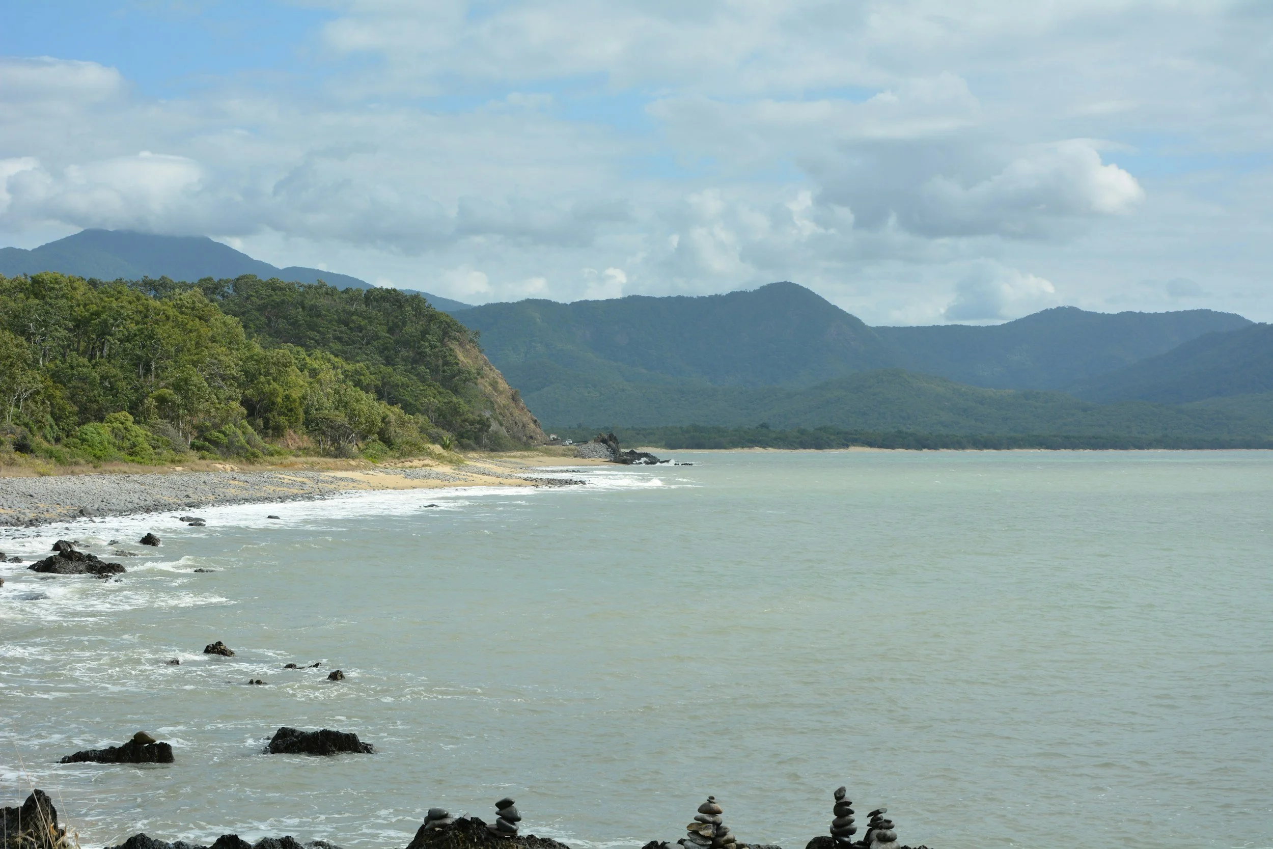 A scenic coastal view with a rocky shoreline, green forested hills, and distant mountains under a partly cloudy sky.