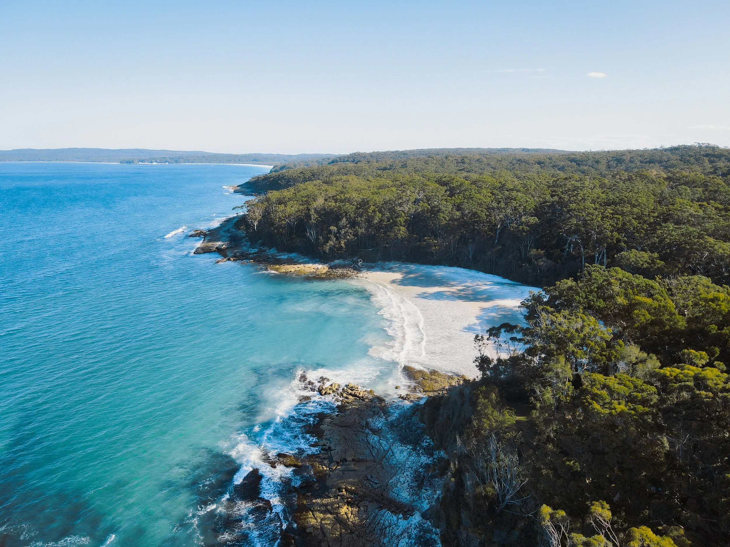 Aerial view of a coastline with clear blue water, rocky shorelines, and dense green trees covering the land.
