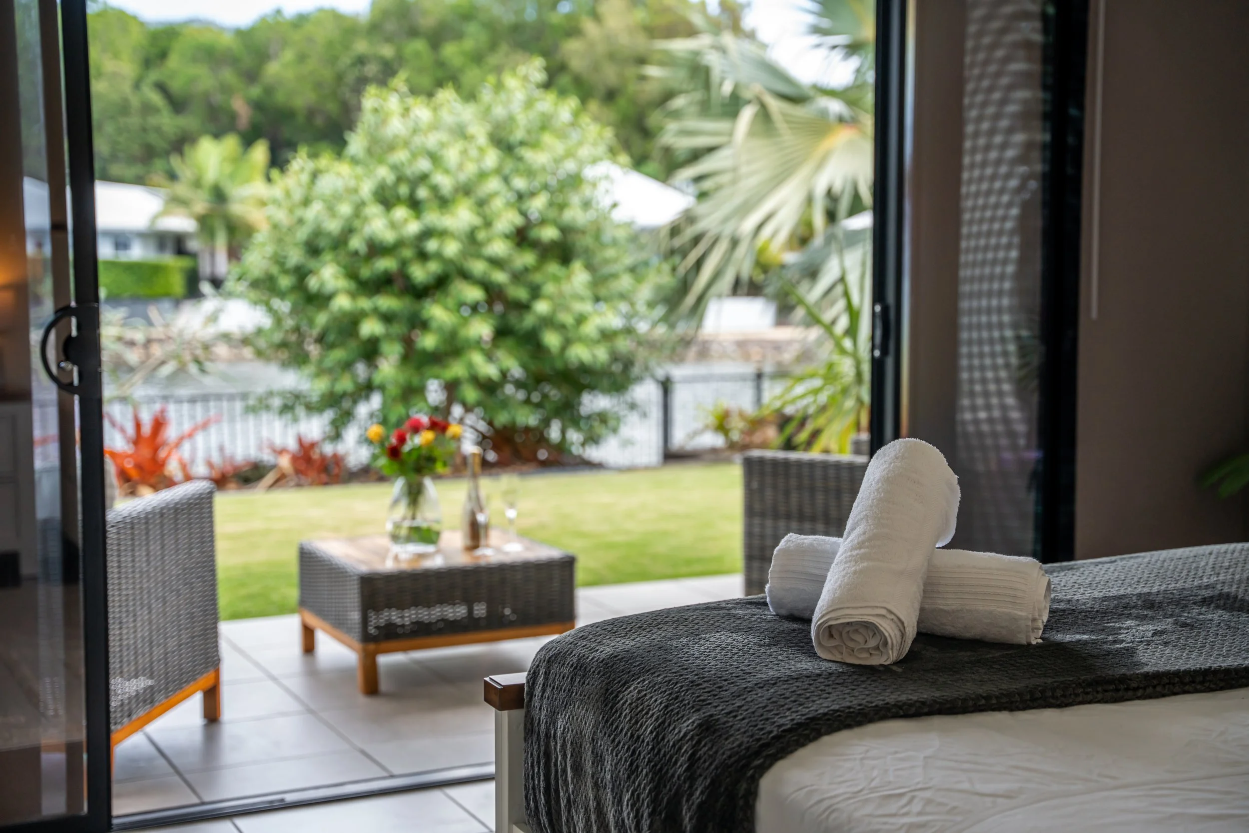 View from inside a room towards a patio with a garden, showing greenery, outdoor furniture, and a table with a vase of flowers.